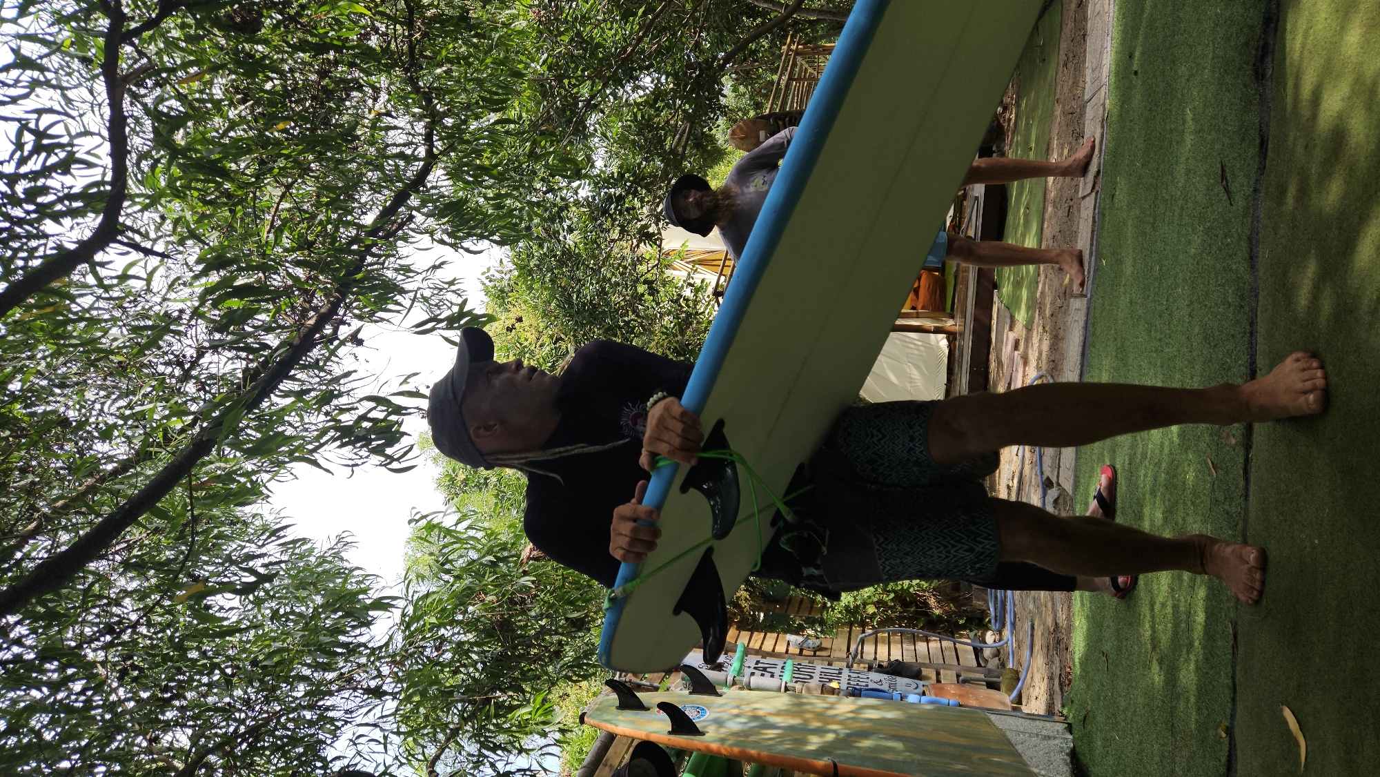 The image, taken outdoors in a green space in what appears to be Cam Lam, Vietnam, shows a man holding a surfboard. The man is wearing a black long-sleeved shirt, patterned shorts, and a baseball cap, and he has dreadlocks. He is carrying a light-colored surfboard with a blue trim and green leash.
Behind him, partially visible, is another person standing on what seems to be artificial turf. The background features lush greenery including trees. In the foreground, another surfboard is visible near a cart.