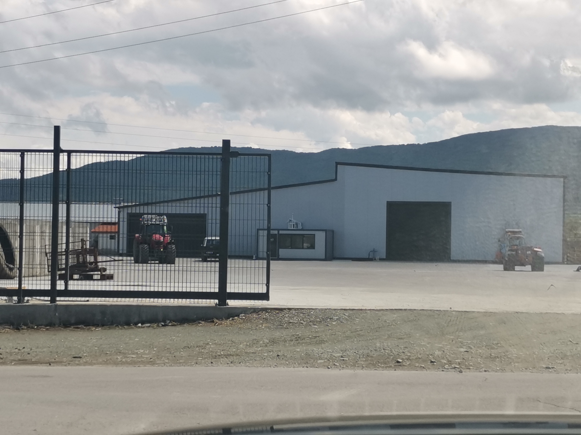 The image depicts an industrial or agricultural facility located in Zhivkovo, Bulgaria, with mountains in the background. The scene is set on an overcast day. In the immediate foreground, a paved road gives way to a gravel shoulder, behind which a black metal fence with an open gate spans across the midground.
Beyond the fence is a large, paved outdoor area. Two modern, light-colored warehouse-style buildings with dark trim are visible. A small, white modular building, possibly a guard booth or office, is situated between the two main buildings.
Within the fenced area, a red tractor with a front loader or attachment is parked to the left, partially obscured by the fence. Another orange-red tractor is visible in the paved area to the far right, facing away from the viewer. Other industrial equipment, including what appears to be a large tire and a metal frame, are near the left side of the fence.
In the background, a range of forested mountains stretches across the horizon under a cloudy sky. Overhead, faint power lines are visible against the clouds. No discernible text is legible on any of the structures or vehicles.