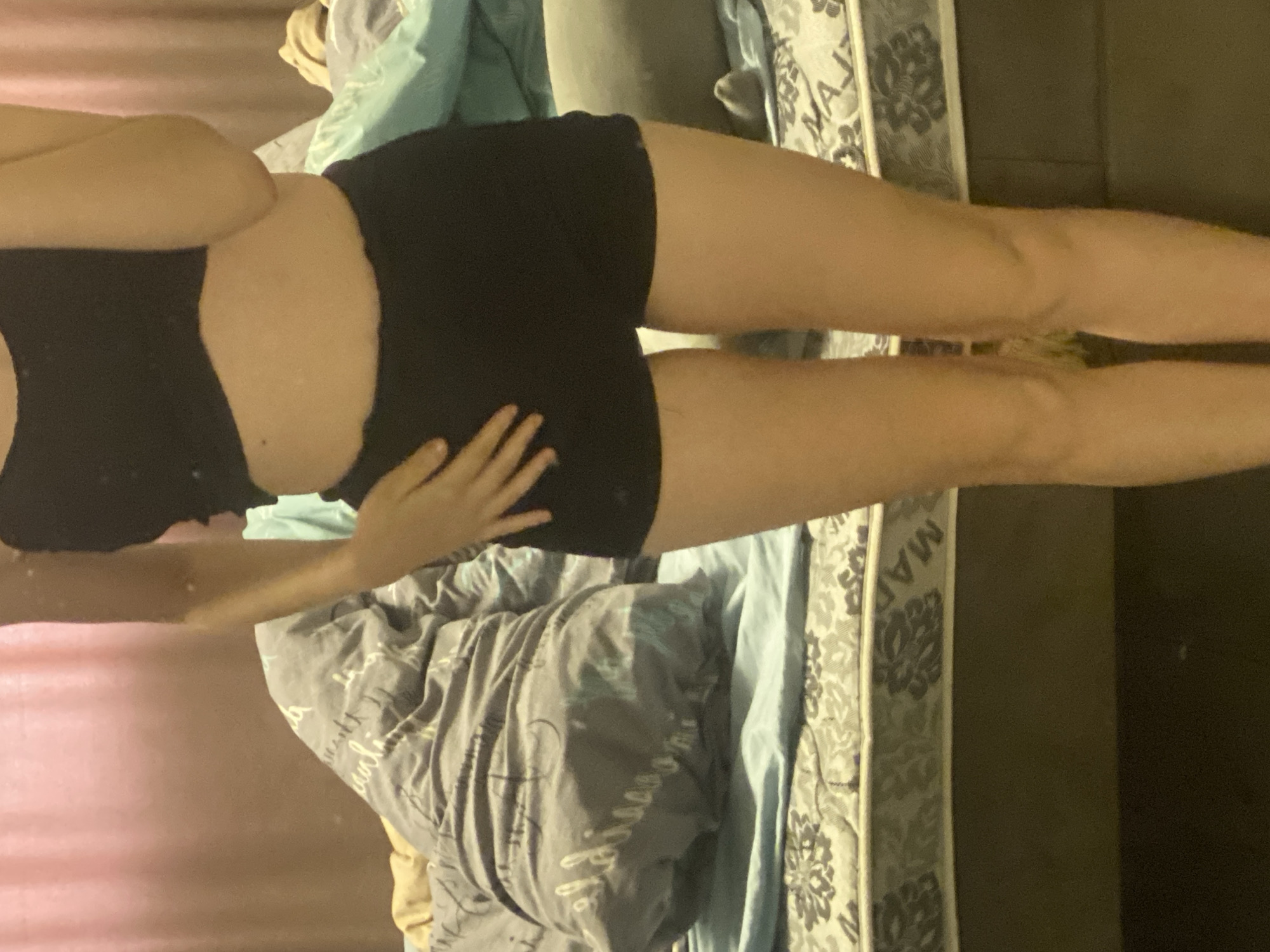 A person wearing a black sports bra and black shorts is positioned on a bed. The bed has a mattress with a floral pattern on its side, and is covered with a wrinkled grey comforter that has a white design on it, and light blue sheets. Part of a hand is visible, resting on the person's hip. The background shows an indistinct interior with a dark wall. The location is not discernible from the image.