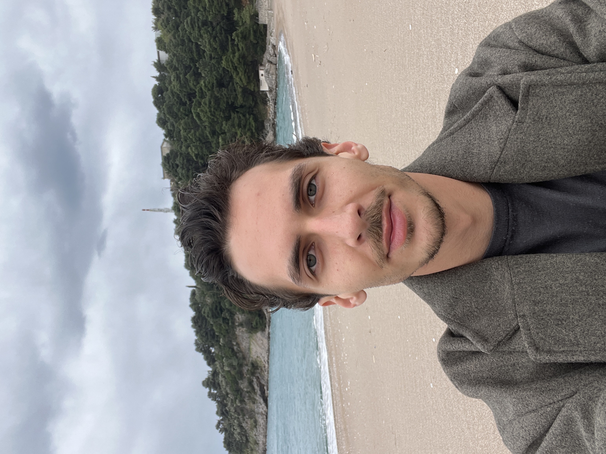 The image is a selfie taken at a beach in Boreti, Montenegro. The subject is a man with dark hair, light eyes, and a trimmed beard, wearing a grey coat over a dark shirt. He is positioned in the foreground. Behind him is a beach with light-colored sand, and the turquoise water of the Adriatic Sea. To the left, the shoreline is lined with dense green vegetation. The sky is overcast. A tall, thin structure is visible amongst the trees in the distance.