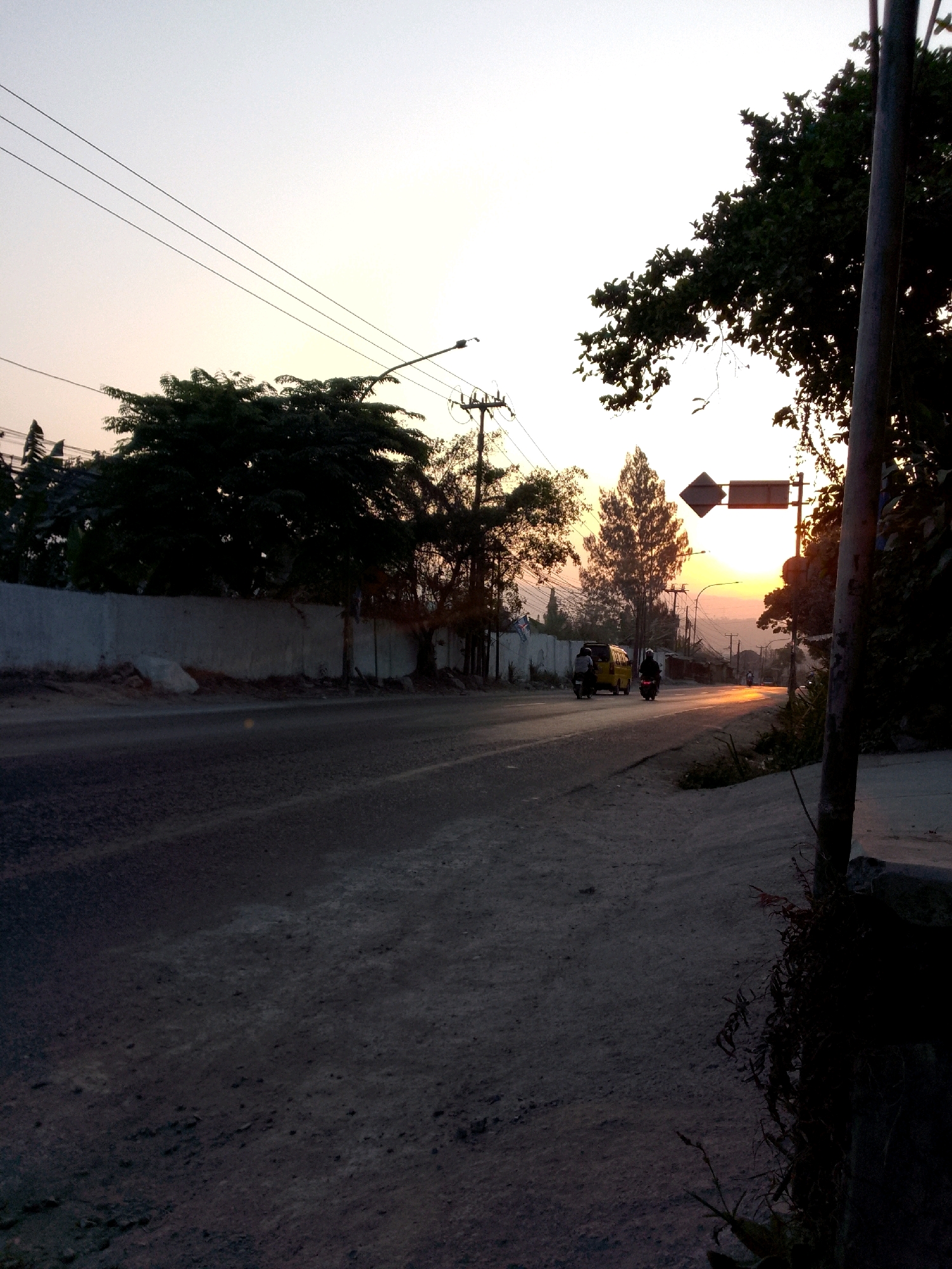 This image captures a roadside scene during sunset in Gunungmasigit, Indonesia. The road is paved and appears to be a main thoroughfare, with a concrete wall and trees lining one side. Several vehicles are visible, including a yellow van and two motorcycles, one of which is being ridden by a person wearing a white helmet. The sun is setting in the background, casting a warm glow over the sky and reflecting on the road surface. A traffic signpost with directional arrows is also present. The overall atmosphere is one of a typical evening commute in a developing urban area.