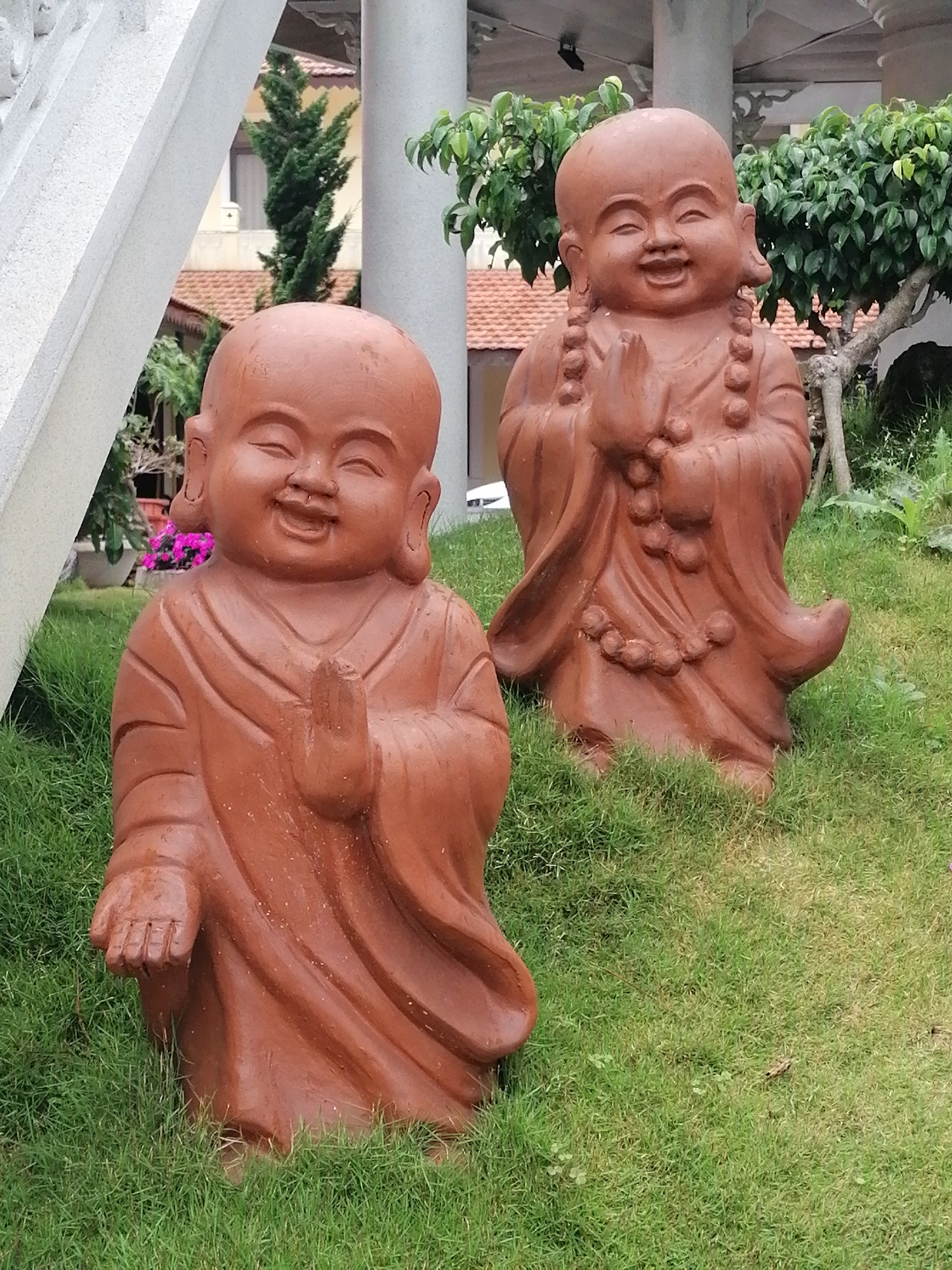 Two terra cotta statues of laughing Buddhist monks are positioned on a lush green lawn in front of a building with a tiled roof and a white column. The monk in the foreground is on the left, facing the camera with its hands in a prayer-like gesture and an open mouth. The second monk, positioned slightly behind and to the right, also has its hands in a similar pose, with a wide smile. Green trees and bushes are visible in the background. The statues appear to be part of a garden or temple. The overall scene is tranquil and decorative.