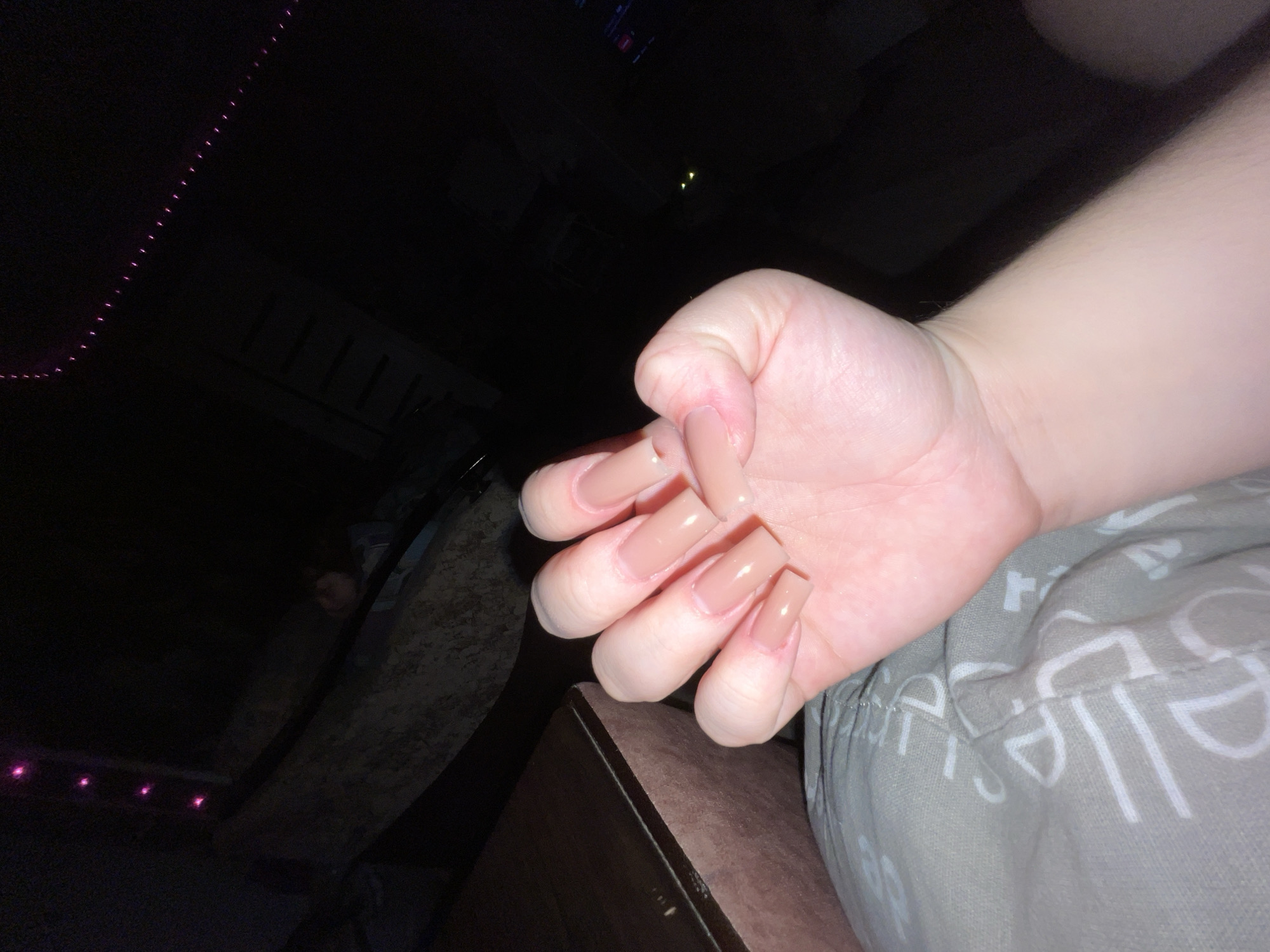 A close-up, low-angle shot depicts a human hand with long, nude-colored acrylic nails. The hand is fair-skinned and appears relaxed, resting on patterned gray and white fabric. The background is dark, with faint pink LED lights visible on the left and vague shapes suggestive of furniture or equipment in the distance. The location in Chaikovsky, Russia is indeterminable from this image.