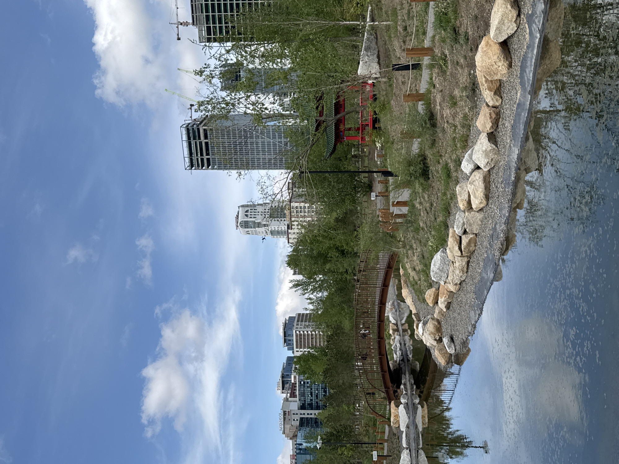 The image depicts a park scene in Ulaanbaatar, Mongolia. A water feature runs along the right side of the frame, lined with rocks. A metal bridge spans the water, connecting to a path lined with wooden posts and rope. Beyond the trees, multiple high-rise buildings stand against a partly cloudy blue sky. A red and green pagoda-style structure is also visible among the trees. Construction cranes are positioned near the taller buildings in the background.