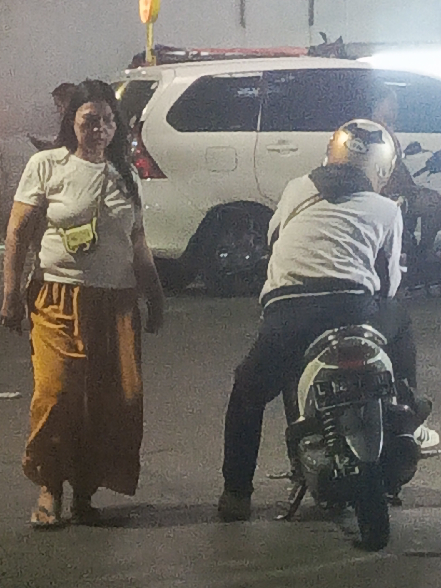 In Surabaya, Indonesia, at night, a woman with dark hair and a white t-shirt stands to the left of the frame, wearing a yellow skirt and sandals. She carries a small, light-colored bag across her chest. To her right, a person wearing a light-colored jacket and dark pants is seated on a motorcycle, their head turned away from the camera, covered by a golden helmet. The motorcycle is parked, and in the background, a white SUV is visible. The scene appears to be on a street, with asphalt visible on the ground.