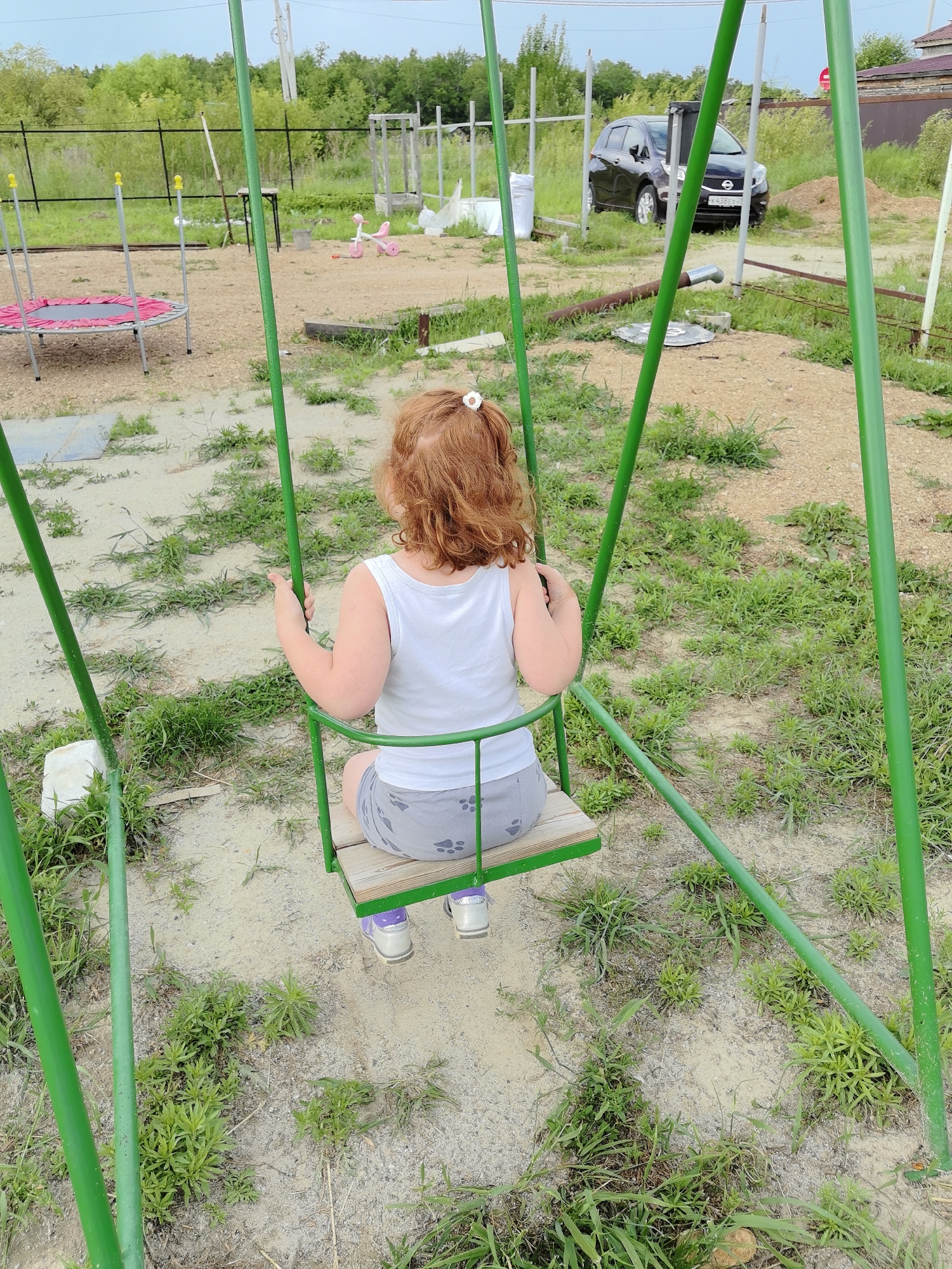 In an outdoor, unpaved area, a young child with curly red hair sits on a wooden swing. The child wears a white tank top and grey shorts with a paw print pattern. The swing set is made of green metal poles. In the background, a trampoline with a pink mat is visible to the left. Further back, a dark-colored car is parked. The environment appears to be a backyard or a rural area, with scattered vegetation and a fence in the distance. The sky is overcast.