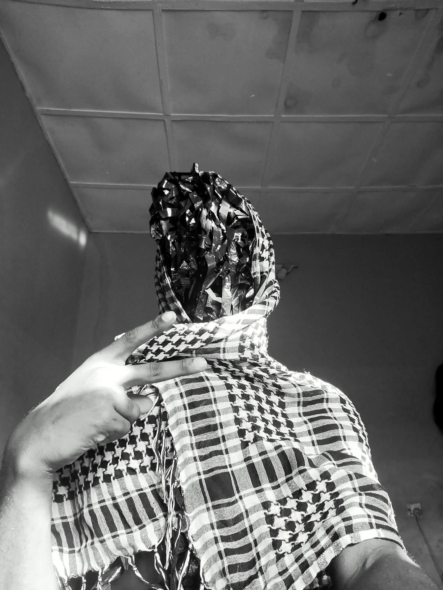 The image is a black and white, low-angle shot of a person whose face is obscured. The head is covered in crumpled, reflective material. A keffiyeh is wrapped around the neck and shoulders. A hand is visible, gesturing with two fingers extended in a peace sign. The background consists of a plain wall with a window on the left side, and a tiled ceiling. The location is a room interior; no details suggest a specific location within the city of Keffi, Nigeria.
