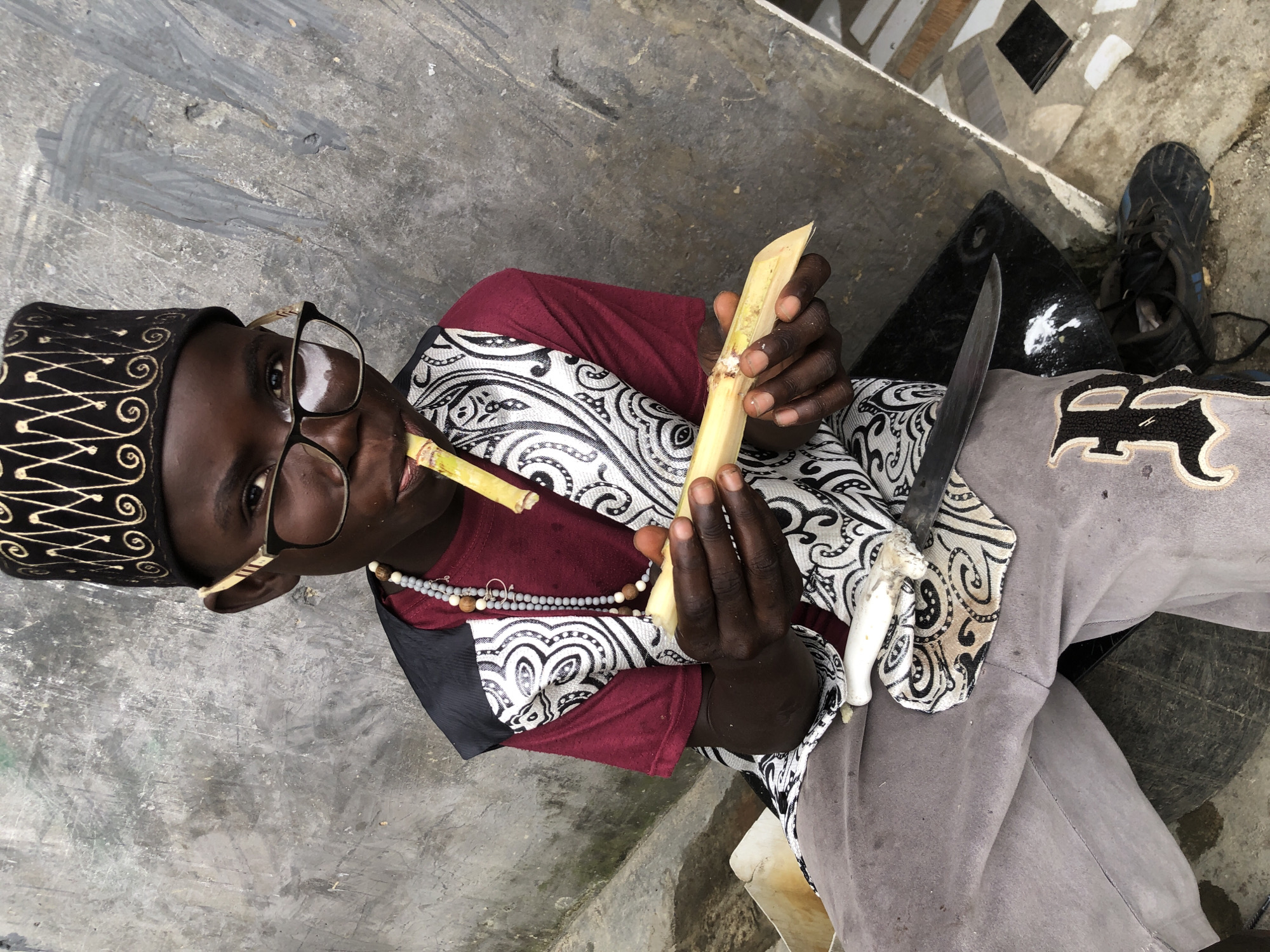 A young Black boy, likely a child, is seated outdoors against a textured gray wall. He wears a maroon t-shirt with a black and white patterned vest over it, gray pants, black shoes, and a black and gold patterned kufi hat. He also wears glasses and a beaded necklace. The boy is holding a piece of sugarcane, with one end near his mouth, and he is in the process of chewing or preparing to chew it. His hands are engaged with the sugarcane, and a knife with a silver blade and a white handle is positioned near his legs, resting on a black mat. The environment suggests a somewhat gritty urban setting in Ado, Nigeria, possibly a backyard or street.