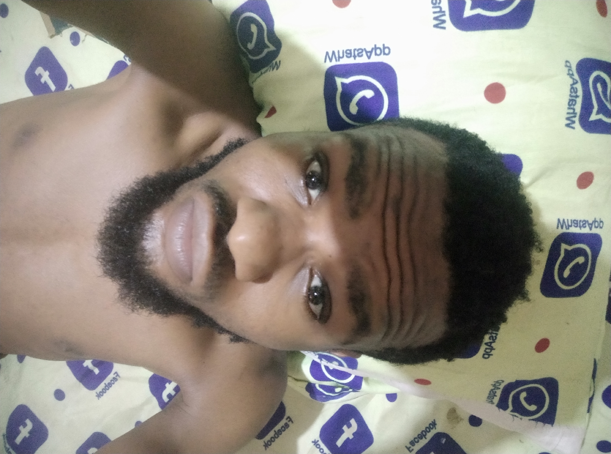The image presents a close-up, slightly tilted view of a dark-skinned man with a full beard, lying down. He looks directly at the camera with a neutral expression. His short, dark, curly hair frames his face, and his forehead shows visible wrinkles. The man appears bare-chested, with his shoulders and upper chest visible in the frame.
The background is a light-colored bedsheet or pillowcase covered in a repeating pattern. This pattern prominently features logos for popular social media applications: the WhatsApp logo (a white phone icon inside a speech bubble on a purple square) and the Facebook logo (a white 'f' on a purple square). The corresponding brand names, "WhatsApp" and "Facebook," are also printed multiple times within the pattern, appearing in various orientations, including mirrored, rotated, and upside down. Small red and purple polka dots are scattered throughout the patterned fabric.
The lighting suggests an indoor setting. No specific activity beyond lying down is discernible, and the time of day, weather, or precise location cannot be determined from the image.