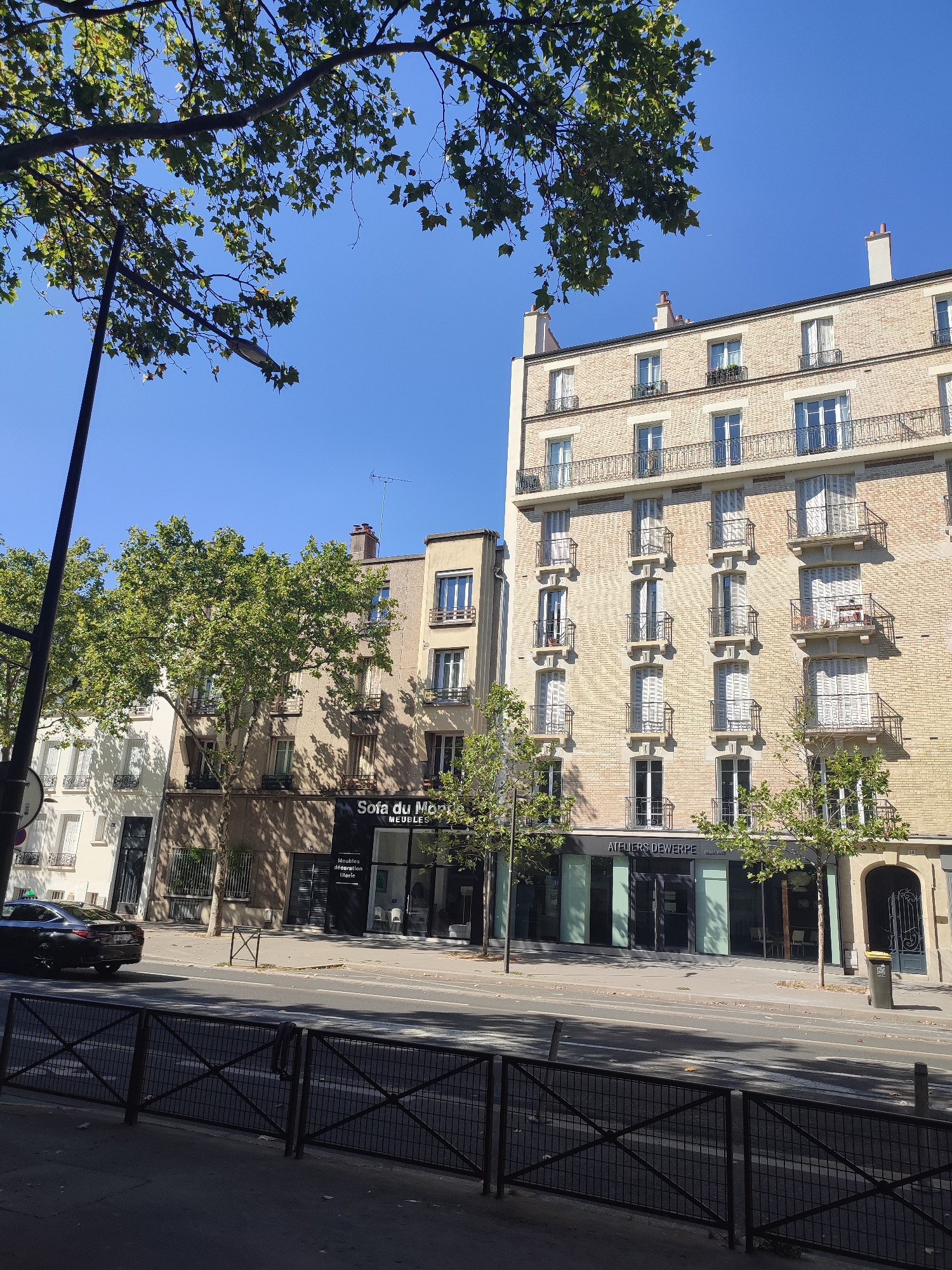 The image captures a street scene in Boulogne-Billancourt, France. A light yellow brick building with multiple stories dominates the right side of the frame, featuring numerous windows, balconies with wrought-iron railings, and dormer windows on the top floor. Adjacent to it, a slightly shorter building of a different color is visible. In the foreground, a street with a pedestrian crossing is bordered by a black metal fence. A dark-colored car is parked on the left side of the street. Trees with green foliage are interspersed along the sidewalk and in front of the buildings, casting shadows on the pavement. The sky is clear and blue, indicating a sunny day. Signage on the storefronts includes "Sofa du Monde MEUBLES" and "ATELIERS DEWERPE." The overall impression is that of a typical urban street in a European city.