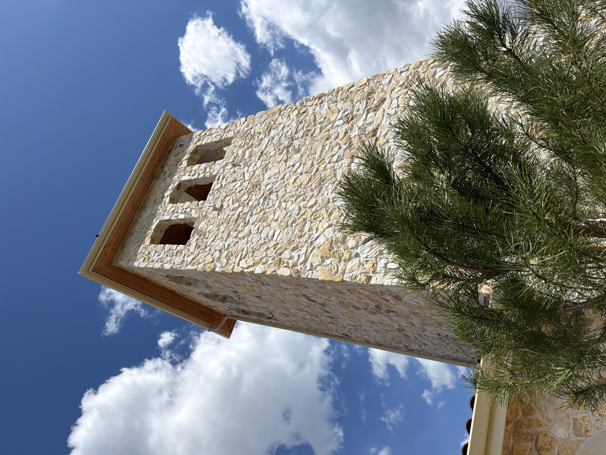 A low-angle shot captures a stone tower situated in Kocherinovo, Bulgaria. The tower, constructed from light-colored, roughly-shaped stones, has three arched openings. A wooden trim outlines the top edge of the tower. A green pine tree partially obscures the tower's right side. The background is a bright blue sky filled with white, puffy clouds.