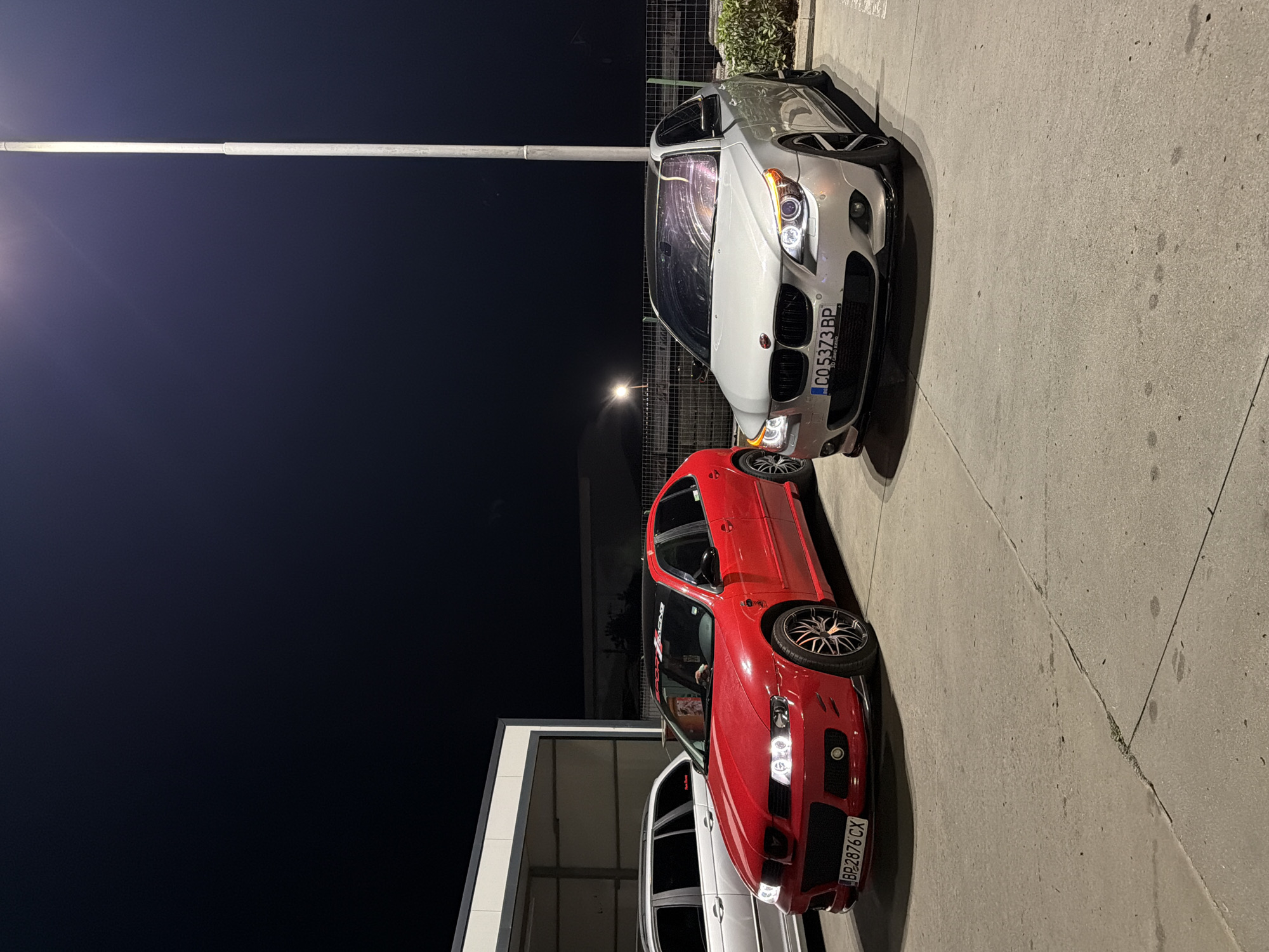 This is a vertical, eye-level, outdoor shot taken at night in Gotse Delchev, Bulgaria. The scene features a row of cars parked on a concrete surface. In the foreground, a bright red car is prominently displayed, its headlights on. Behind it, a silver car is parked, also with its headlights illuminated. A third car, mostly obscured, is visible beneath the red car. The image captures a night scene, likely after dark, given the illumination from the car headlights and the dark sky. The concrete ground suggests an urban or semi-urban setting, possibly a parking lot or a street. The presence of multiple cars indicates a gathering or a simple parking arrangement. No people are visible in the image. Visible text includes license plates on the red car, reading "BP2876 CX", and on the silver car, reading "C05373BP". The overall mood is neutral, focusing on the cars and the nighttime environment.