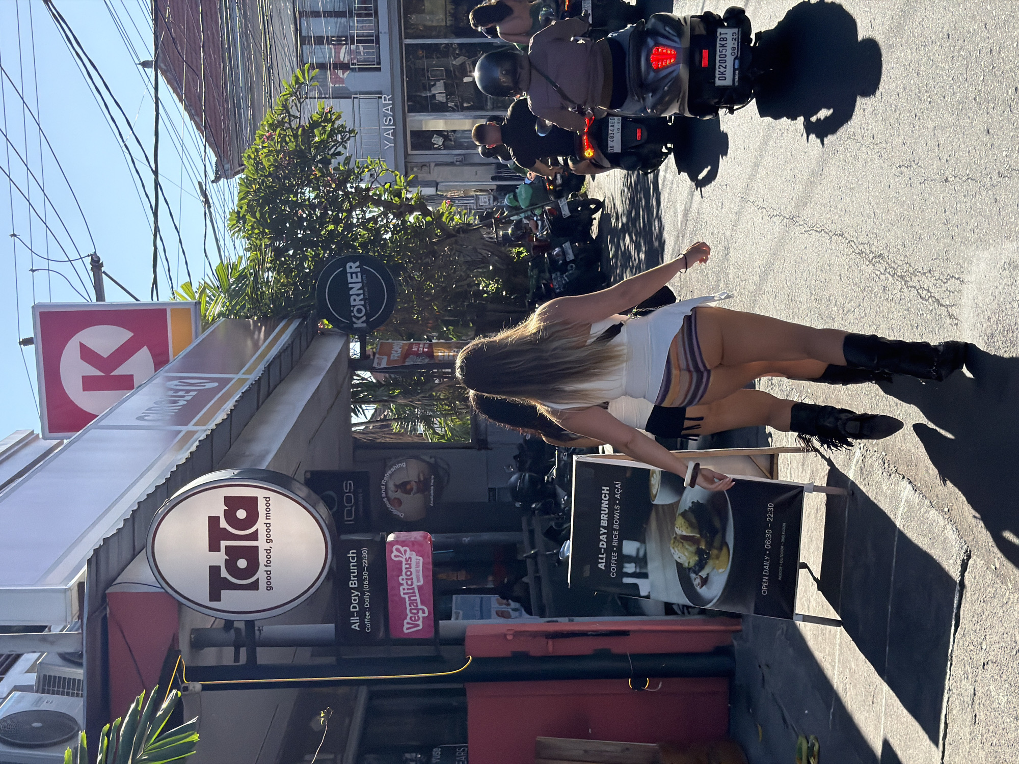 The image shows a street scene in Mengwi, Indonesia, featuring a street and surrounding businesses. In the foreground, a woman in a white top, striped shorts, and black boots stands next to a sandwich board advertising "ALL-DAY BRUNCH" at a place named "Tata" (advertised as "good food, good mood"). Adjacent to Tata is a Circle K convenience store and a sign for "Veganlicious." A sign for "Körner" is also visible. Motorbikes are parked and driving along the street, with riders wearing helmets. A building labeled "YASAR" is in the background. The image is taken during daylight, with strong sunlight casting shadows on the pavement.