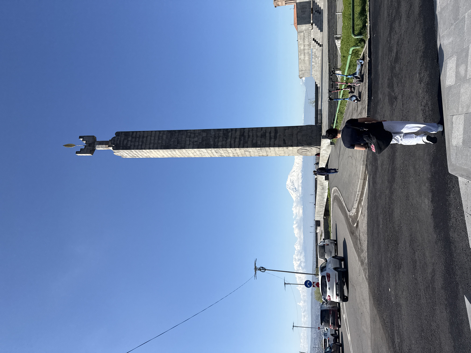 The image shows the Yerevan Cascade monument in Yerevan, Armenia, on a clear, sunny day. The monument is a large, tiered structure with a central obelisk topped with an eternal flame. In the background, there is the snow-capped Mount Ararat visible in the distance. People are walking near the monument. There are cars parked along the street in front of the monument. There are also electric scooters visible next to the steps.