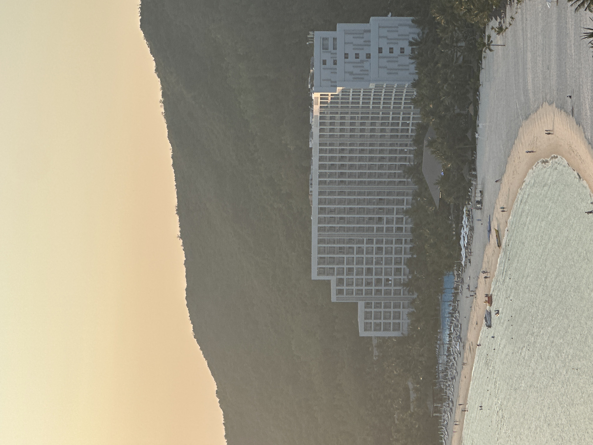 The image presents an elevated view of a coastal resort scene, characterized by soft, warm light suggesting either early morning or late afternoon. A large, multi-story white building, likely a hotel or resort, dominates the center-right. It features a modern, terraced architectural style with numerous windows, and is nestled at the foot of a sprawling, tree-covered mountain or hillside.
To the right of the building and mountain lies a wide, sandy beach that gently curves around a calm bay or ocean inlet. Palm trees are visible along the border between the resort property and the beach. The beach is equipped with numerous lounge chairs and umbrellas, some distinctly blue, indicating a leisure area. Several small figures of people are scattered along the shoreline and in the shallow, clear water, engaged in activities such as walking or relaxing. Small boats or kayaks and buoys are also visible near the water's edge.
The mountain range on the left is densely covered in lush green foliage, fading into a gentle atmospheric haze towards the background. The sky, visible to the far left, exhibits a soft, yellowish-orange hue consistent with a sunrise or sunset. The overall impression is one of a peaceful, tropical vacation destination. No discernible text is visible in the image.