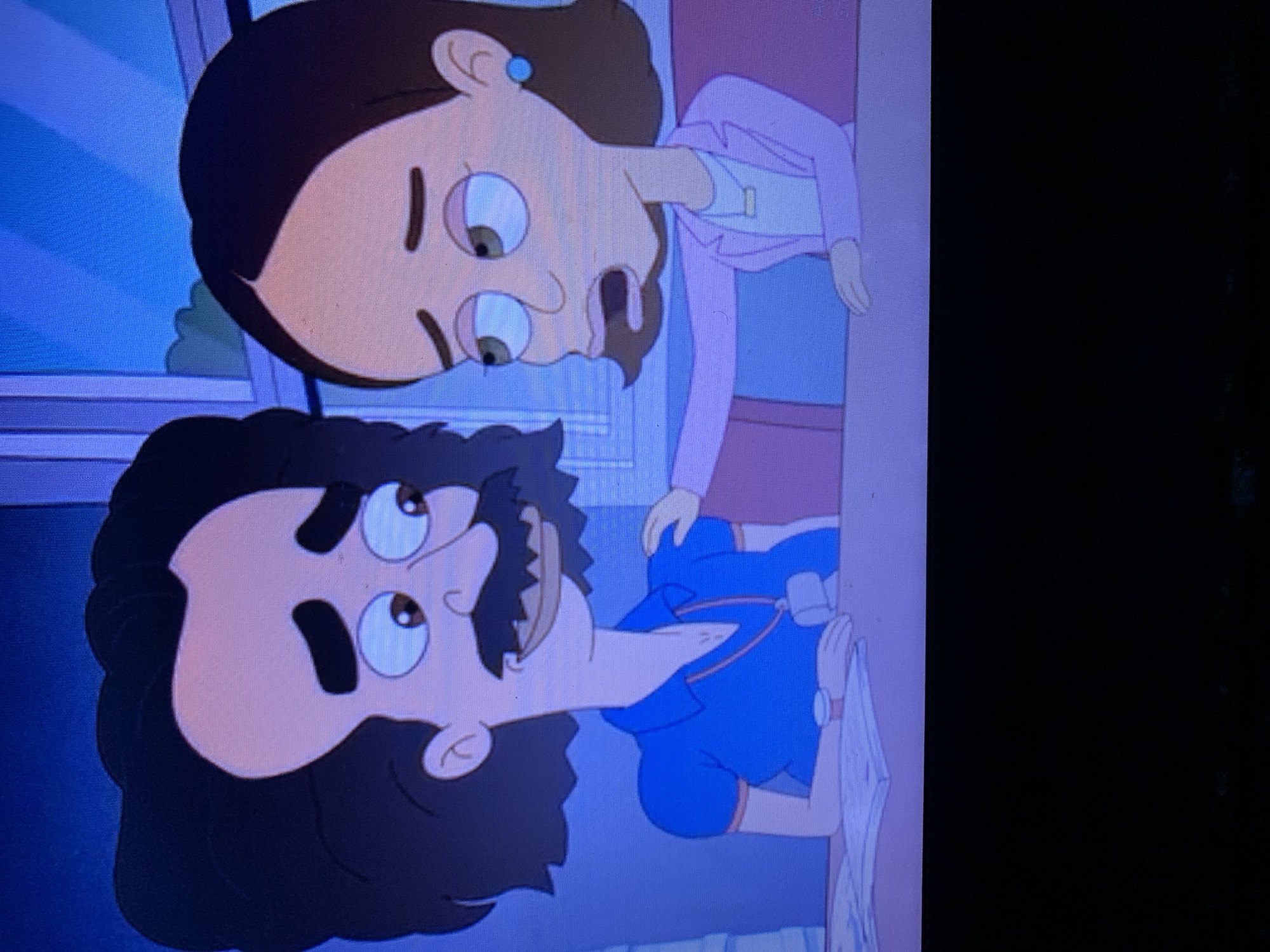 The image shows a close-up, animated depiction of two characters, a man and a woman, sitting at a table. The man, on the left, has dark, curly hair, a prominent mustache, and is wearing a blue polo shirt. He has his hand resting on the table. The woman, on the right, has dark hair pulled back and is wearing a pink blazer over a light-colored shirt. Her left hand is resting on the man's shoulder, and her right hand is also on the table. Both characters appear to be looking off to the side with concerned or troubled expressions. The background is blurred and appears to be an indoor setting, possibly an office or classroom, with a window visible. There is no information in the image that places the scene in Jos, Nigeria.