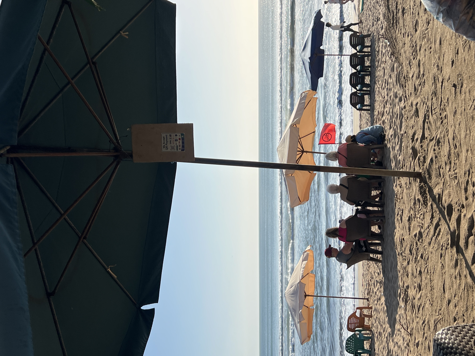 The image depicts a scene on Kuta Beach, Indonesia, with several people enjoying the ocean view. In the foreground, a large dark umbrella provides shade, with a sign attached to its pole indicating "We Accept QR Payment." Several beach chairs and small tables are set up on the sand.
Four individuals are seated at a table under an orange umbrella, facing the ocean. To the left, two empty green and brown plastic chairs are visible. To the right, a group of four empty black plastic chairs are arranged, and further right, a person walks along the shoreline under a large blue umbrella. The ocean has moderate waves, and the sky is clear and blue. The overall atmosphere suggests a relaxed beachside setting.