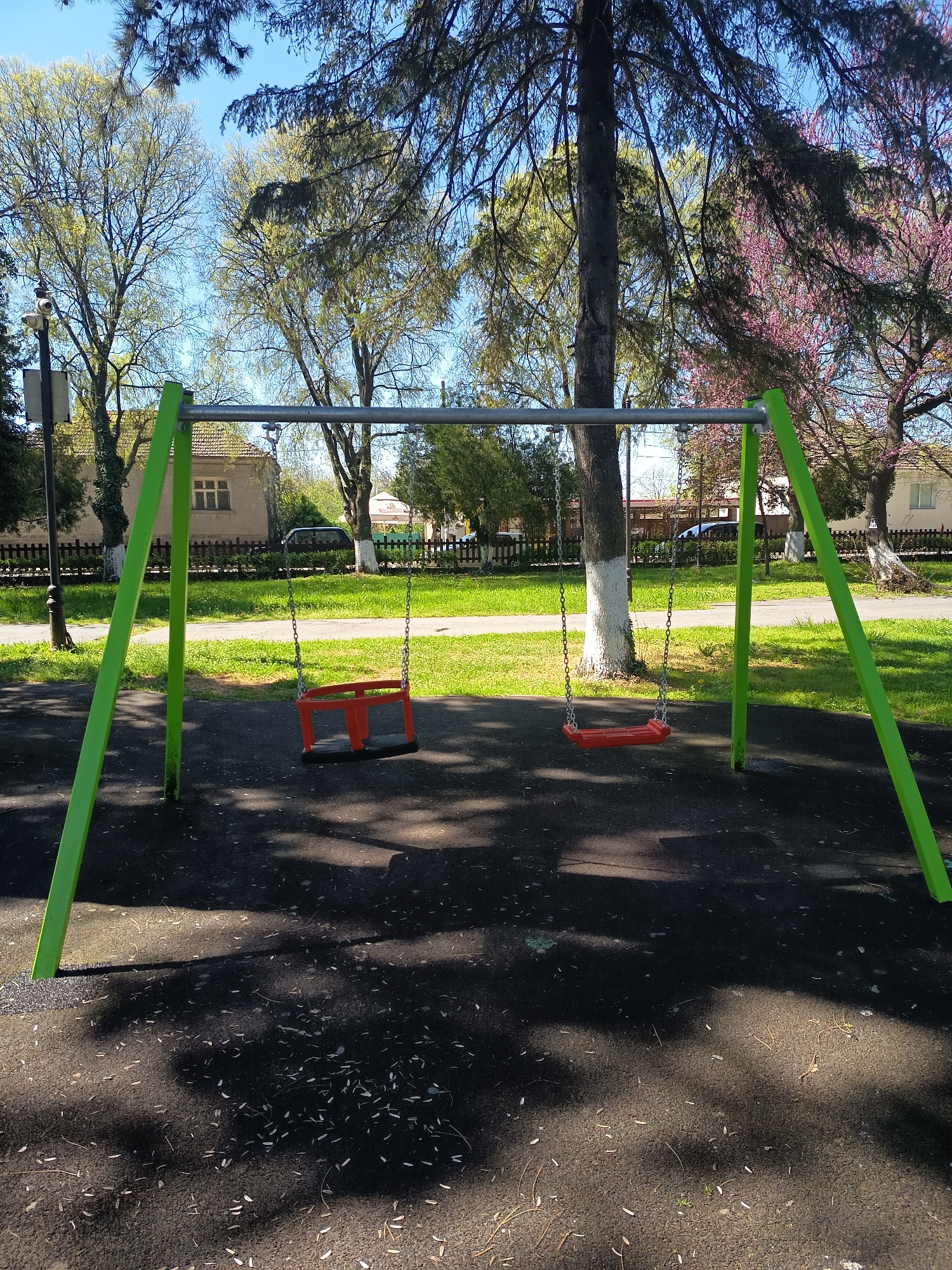The image depicts a swing set in a park, likely located in Sungurlare, Bulgaria. The swing set has a metal frame with green painted supports and two swings, one a typical seat and the other a bucket style, both suspended by chains. The ground beneath the swings is a dark, rubberized surface covered in scattered leaves. In the background, there is a grassy area, trees, and buildings, one with a light beige exterior and dark trim. The sky is blue and clear. A tree trunk, painted white at its base, is located directly behind the swing set. No people are visible in the image. The scene is well-lit, suggesting it is daytime.
