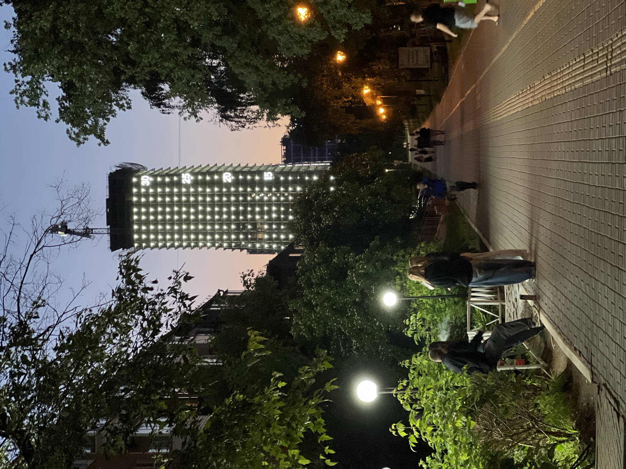 The image depicts an evening scene along a paved walkway in Izhevsk, Russia. A tall, unfinished skyscraper dominates the background, its facade illuminated with numerous bright lights, possibly indicating construction progress or decorative lighting, with visible numbers "30", "25", and "20" on its side. The sky is a twilight gradient of blue and orange. To the left of the walkway, lush green trees and bushes line the path, with two modern, spherical streetlights casting a warm glow. A few people are visible on the walkway. In the foreground, two individuals, one seated on a low barrier and smoking, the other standing, are silhouetted against the ambient light. Further down the path, a group of people are walking. The walkway itself is paved with brick-like tiles. The overall atmosphere is one of urban twilight, with a blend of natural greenery and architectural development.
