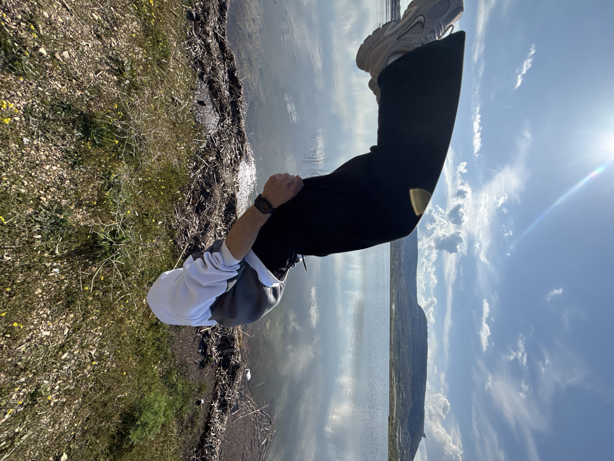 A person in black pants and light-colored sneakers is standing near a body of water. The person is also wearing a gray and white hooded sweatshirt and a watch on their left wrist. To the left of the person is a grassy area with some yellow flowers. The water reflects the sky, which is blue with some clouds. In the distance, there is a mountain range. The location is likely near a lake or reservoir outside of Shumen, Bulgaria, given the landscape.