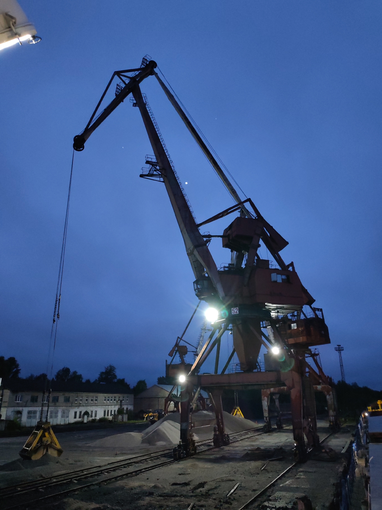 This image depicts an industrial port or yard at dusk or night, characterized by a deep blue sky and illuminated by artificial lights. A large, rusty gantry crane dominates the scene, its intricate structure and control cabins highlighted by several bright floodlights. The number "52" is visible on the side of this central crane.
To the left, another crane's boom extends, suspending a large, yellow clam-shell grab bucket poised above a substantial pile of light-colored aggregate material, likely sand or gravel. Multiple similar piles are scattered across the ground. A network of railway tracks runs across the foreground and mid-ground, indicating a transport hub for bulk goods. In the distance on the left, a long, multi-story industrial building with numerous windows appears weathered. A yellow loader vehicle can be seen in the background near the material piles, and a slender tower stands against the sky on the far right. The lower right corner reveals part of a ship or dockside structure.
The clear, dark blue sky features a faint crescent moon or bright star visible near the top of the main crane's structure. The artificial lighting creates an operational atmosphere, illuminating the heavy machinery and the industrial environment. No people are explicitly visible. The scene suggests active loading or unloading operations of raw materials in this industrial setting, likely a port facility in Medvezhyegorsk, Russia.