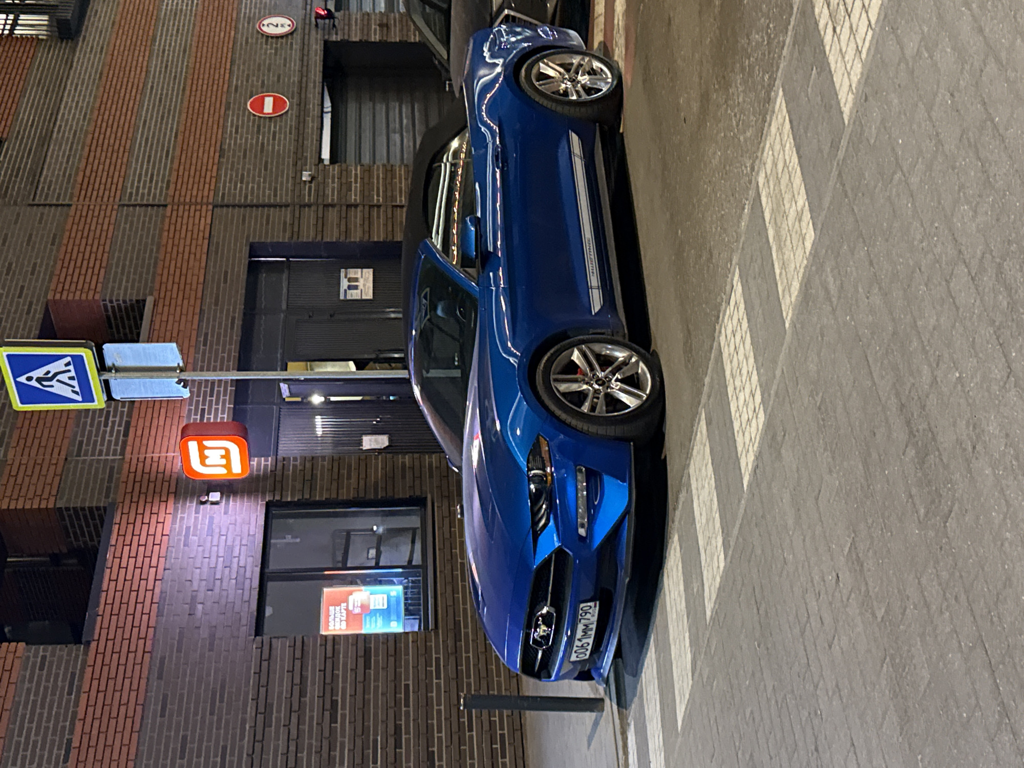 The image captures a blue convertible Ford Mustang parked on a crosswalk in Khimki, Russia. The vehicle, with a license plate reading "O061MM790", is oriented parallel to a building featuring a brick facade with vertical stripes of darker bricks. A lit sign with an orange logo is visible on the building, near a pedestrian crossing sign. Traffic signs indicating a speed limit of 20 km/h and no entry are also present above a commercial space with closed metal shutters. The environment appears to be an urban setting with street lighting.