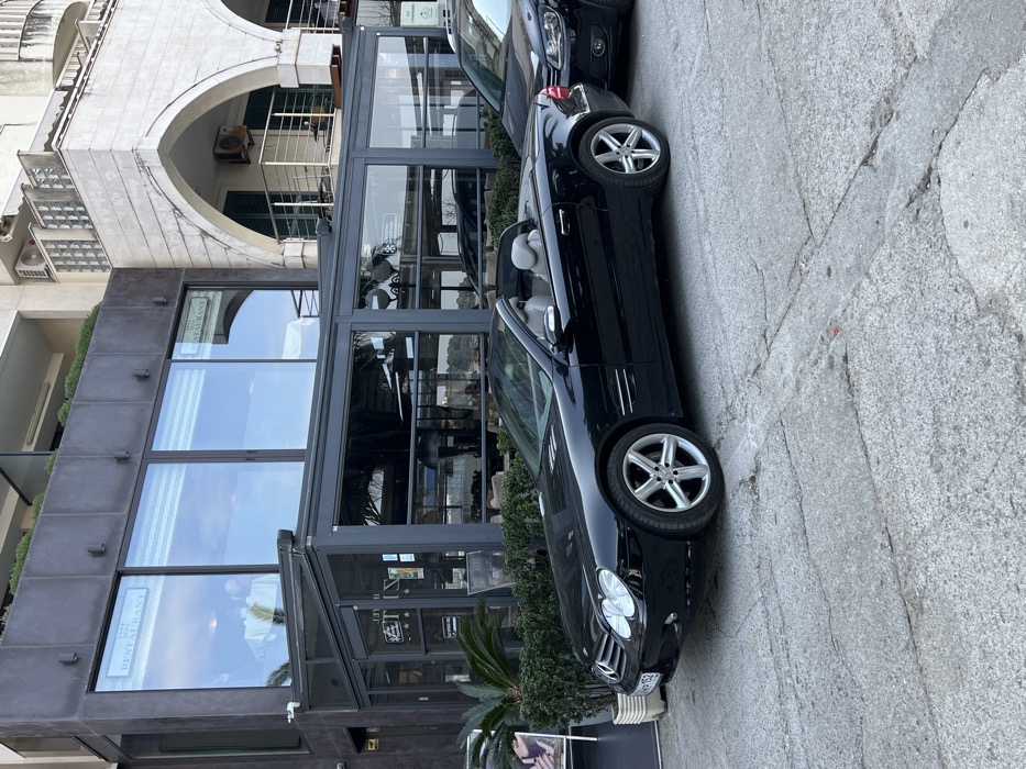 The image depicts a street scene in Budva, Montenegro, featuring a modern restaurant building and an adjacent multi-story structure. In the foreground, a light-colored, cracked asphalt or concrete street surface is visible.
Two vehicles are parked parallel to the buildings. A black Mercedes-Benz SL-Class convertible (R230 generation), with its top down and light grey interior seats visible, is parked prominently. Its front license plate displays "BD-MP-152". Immediately behind the Mercedes, a dark-colored SUV, possibly a Volkswagen, is partially visible.
The building on the left is a contemporary structure with large, dark-framed glass windows on its upper level, where signs indicating "RESTAURANT" are affixed. Below these windows, a glass-enclosed outdoor seating area or conservatory extends forward, also with a dark frame. Green shrubs and potted palm plants are arranged along the front of this area. A white promotional stand is positioned near the front left of the restaurant, next to the plants.
To the right, an older, multi-story building with light-colored masonry features prominent arched balconies with railings. Air conditioning units are mounted on its exterior walls. The sky, reflected in the restaurant windows, appears overcast or hazy. No individuals are visible in the scene.
