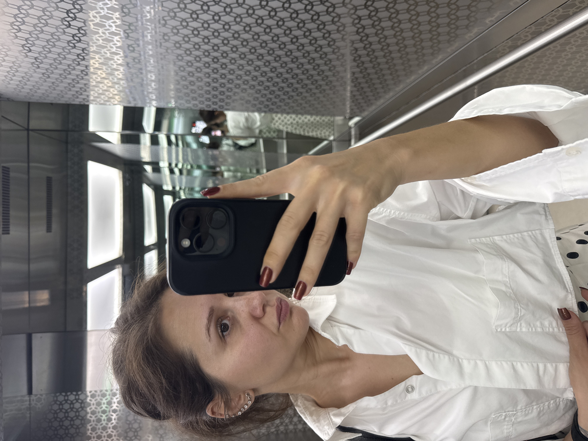 A young woman is depicted taking a selfie with a black smartphone, likely an iPhone, in what appears to be an elevator or a modern mirrored room. The image is oriented vertically, with the woman looking directly towards the camera, holding the phone with her right hand. Her fingernails are painted a deep red. She has medium-length brown hair pulled back or to the side, and a neutral expression with a slight pout. She is wearing a crisp white collared shirt, and a decorative ear cuff or multiple earrings are visible on her left ear.
The background shows highly reflective surfaces. The ceiling features a patterned silver-grey surface with interconnected geometric shapes, possibly circles or hexagons. The mirrored walls reflect internal elements like bright vertical light panels or windows, as well as distorted reflections of the woman and her surroundings. Metallic trims are visible along the edges of the panels. Part of a polka-dot patterned garment is faintly visible in the reflection or beneath her white shirt. The overall setting suggests a sleek, contemporary interior. No specific time of day or external weather can be determined. The provided context indicates the location is Dubai, United Arab Emirates. There is no visible text in the image.