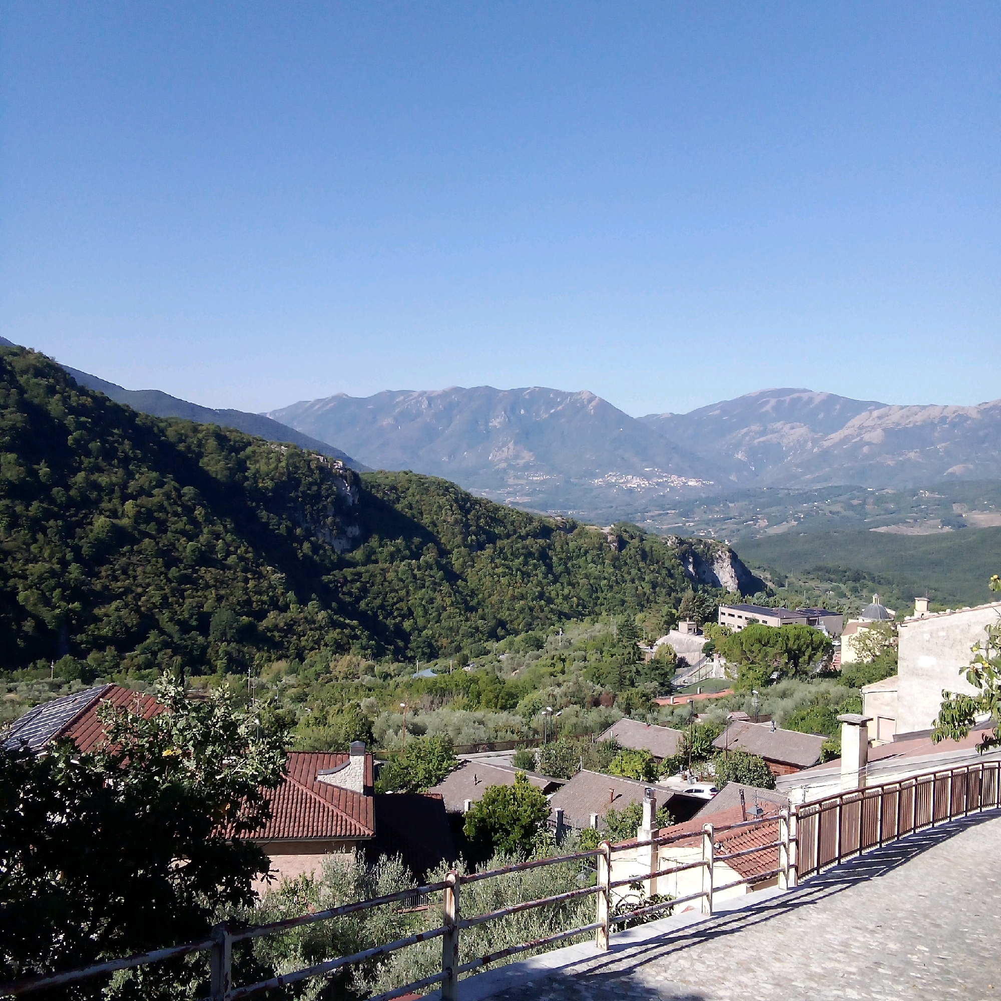 The image shows a panoramic view of the town of Santomenna, Italy, nestled within a valley surrounded by rolling hills and mountains. The foreground is dominated by a cobblestone pathway and a rusty metal railing, casting long shadows from the sun. Beyond the railing, a cluster of houses with terracotta tiled roofs is visible, some featuring solar panels.
In the mid-ground, the landscape transitions to a dense forest covering the lower slopes of the hills. The trees are a vibrant green, indicating a temperate climate. Further into the distance, the valley opens up, revealing more scattered houses and buildings, and a glimpse of a small town or village perched on a distant hillside.
The background is a dramatic mountain range with rugged peaks, partially obscured by a light haze. The sky is a clear, bright blue, suggesting a sunny day. The overall impression is one of a peaceful, rural setting in the Italian countryside. There are no people or animals visible in the frame. The scene is static and serene.