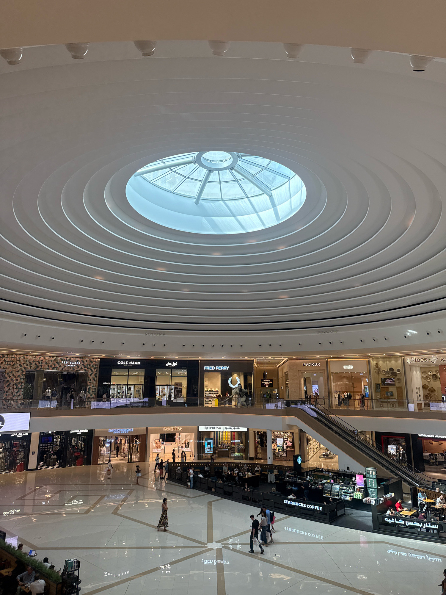 The image shows the interior of a large, modern shopping mall in Dubai, United Arab Emirates. The architecture is characterized by a circular, multi-layered ceiling with a central skylight, allowing natural light to illuminate the space. Suspended from the ceiling are concentric circular elements, creating a visually striking effect.
Below the ceiling, multiple levels of retail stores are visible. Some of the store names that can be discerned include "TED BAKER LONDON", "COLE HAAN NEW YORK", "FRED PERRY", "SANDRO", "maje", "tip toes", "pierre cardin", "EMPORIO ARMANI", and "PANDORA". The stores are designed with glass facades, allowing glimpses of merchandise and displays inside.
The mall floor is polished and reflective, mirroring the lights and architecture above. People are seen walking, browsing, and interacting in the open spaces. An escalator system is visible, connecting different levels of the mall. A "STARBUCKS COFFEE" outlet with seating areas is prominently featured, with customers seated and ordering. The overall atmosphere suggests a busy and upscale shopping environment. The reflection on the floor shows upside down text and patterns.