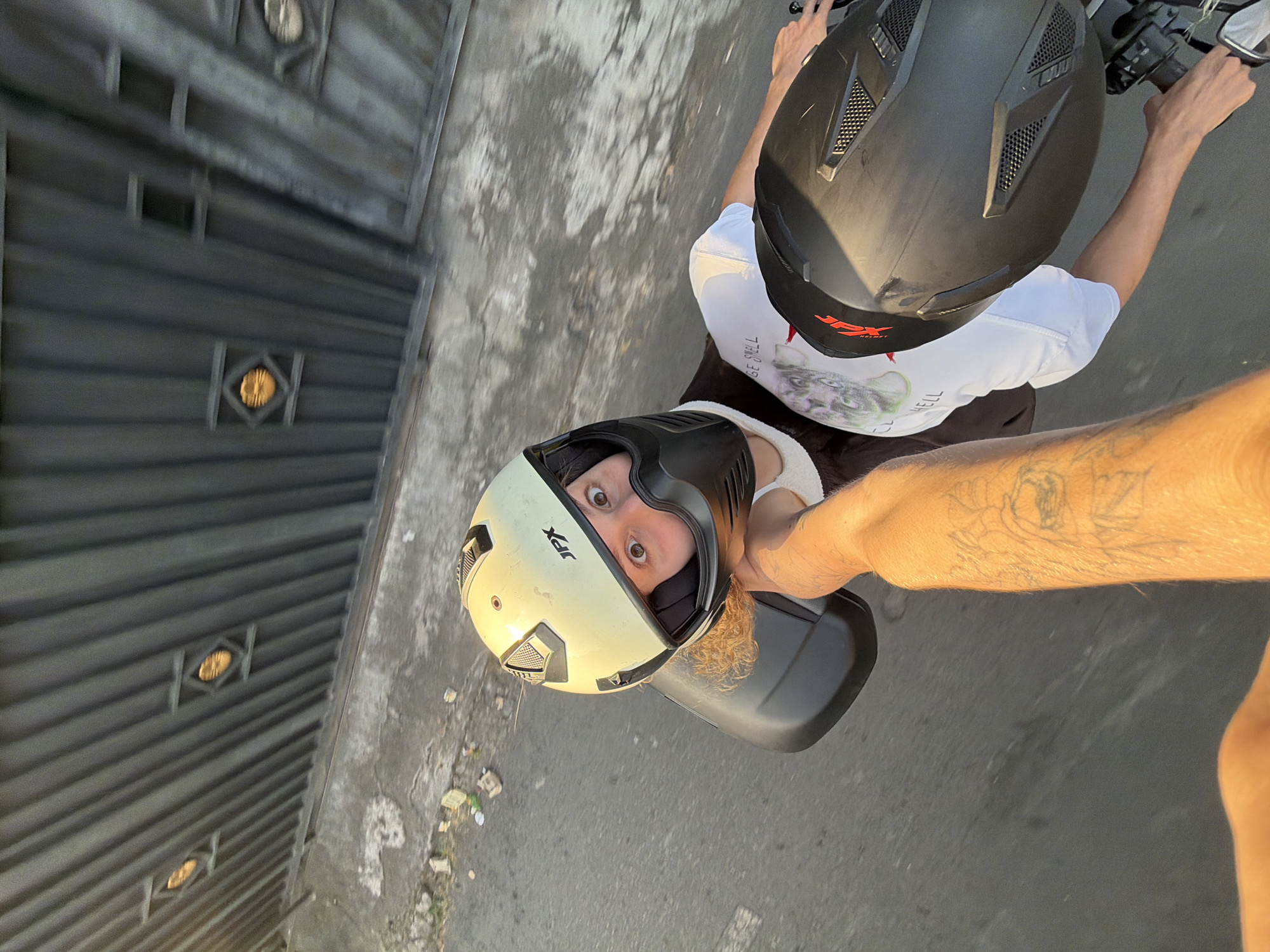 The image shows a first-person perspective of a person riding a motorcycle in Mengwi, Indonesia. The rider is wearing a light-colored helmet with a black chin guard and a white t-shirt with a graphic print. Another person, also wearing a black helmet, is seated behind the rider, holding onto the rider's arm. The background consists of a dark metal gate with diamond-shaped cutouts, a concrete sidewalk with some debris, and a road. The lighting suggests it is daytime, possibly late afternoon due to the warm tones.