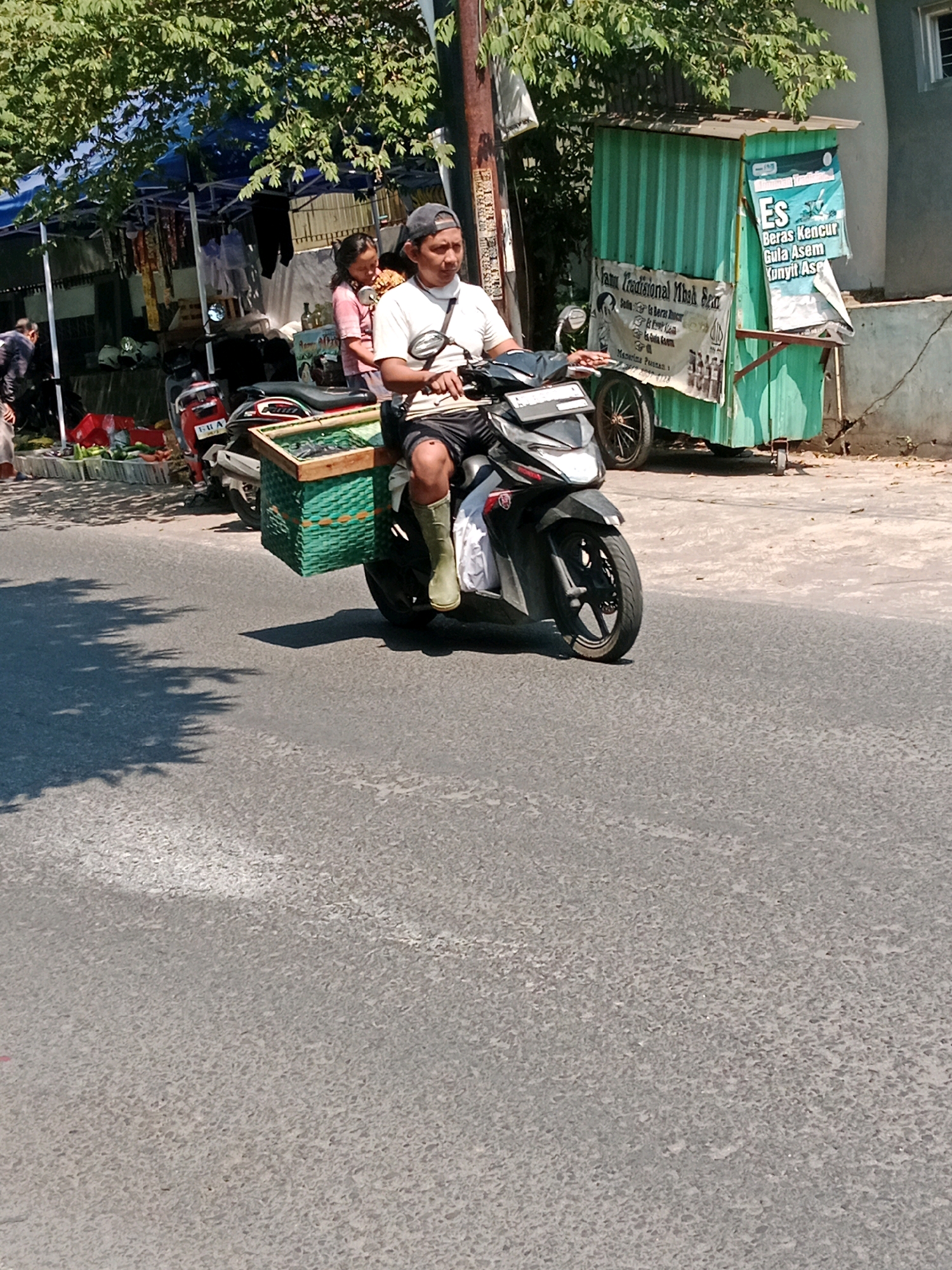 In the city of Banaran, Indonesia, a man is riding a motorcycle on a paved road. He is wearing a white t-shirt, black shorts, a black baseball cap, and green boots. A young girl is sitting behind him on the motorcycle, wearing a pink shirt and a pink hair accessory. Attached to the back of the motorcycle is a green woven basket filled with produce, possibly vegetables. In the background, there is a green food stall with signs advertising drinks. The area appears to be a marketplace, with tents and other stalls visible. Sunlight casts shadows on the road from the surrounding trees and structures.