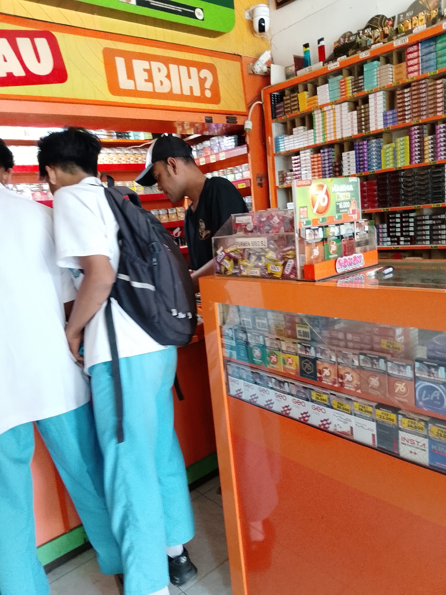 The image captures a street-level view of a convenience store in Pasuruan, Indonesia. The store interior is filled with shelves stocked with various products, primarily cigarettes, displayed behind a vibrant orange counter. A man wearing a black t-shirt and a black and white cap is visible behind the counter, seemingly attending to customers. Two young men, possibly students based on their uniforms, are standing in front of the counter. One is wearing a white t-shirt and blue pants and carrying a black backpack. The other, partially visible, is wearing a white t-shirt and blue pants. The signage above the counter features the words "AU LEBIH?" in bold letters, suggesting a promotional offer or slogan. The overall scene depicts a typical retail environment in Indonesia.