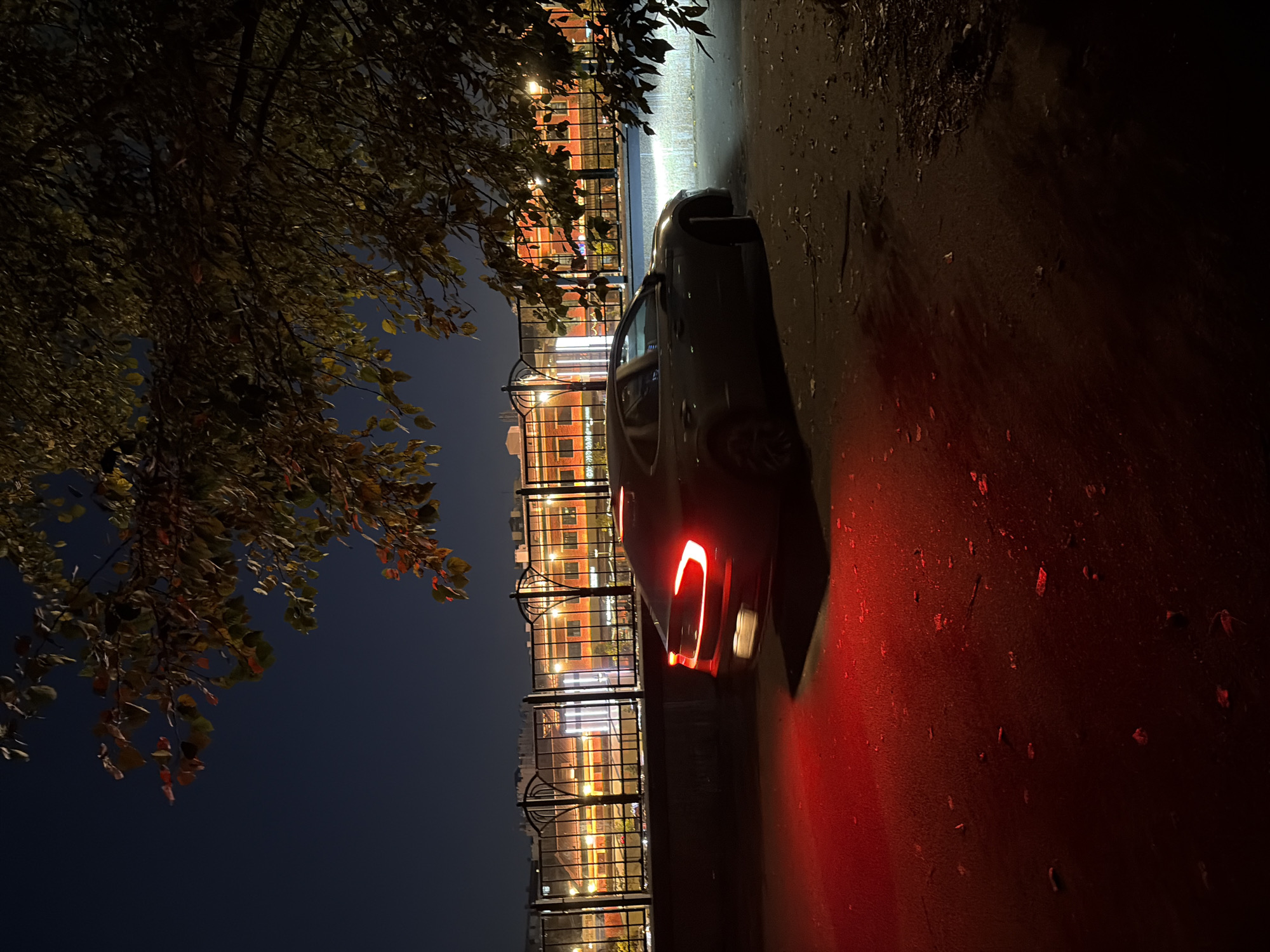 A dark sedan is parked on a paved surface at night. The car's taillights are illuminated, casting a red glow on the ground. The vehicle is positioned in front of a low-lying concrete barrier, behind which is a black metal fence. Beyond the fence, a building with multiple windows is visible, illuminated by warm interior lights. Trees with visible leaves overhang the scene from the top. The overall impression is a quiet urban setting at night.