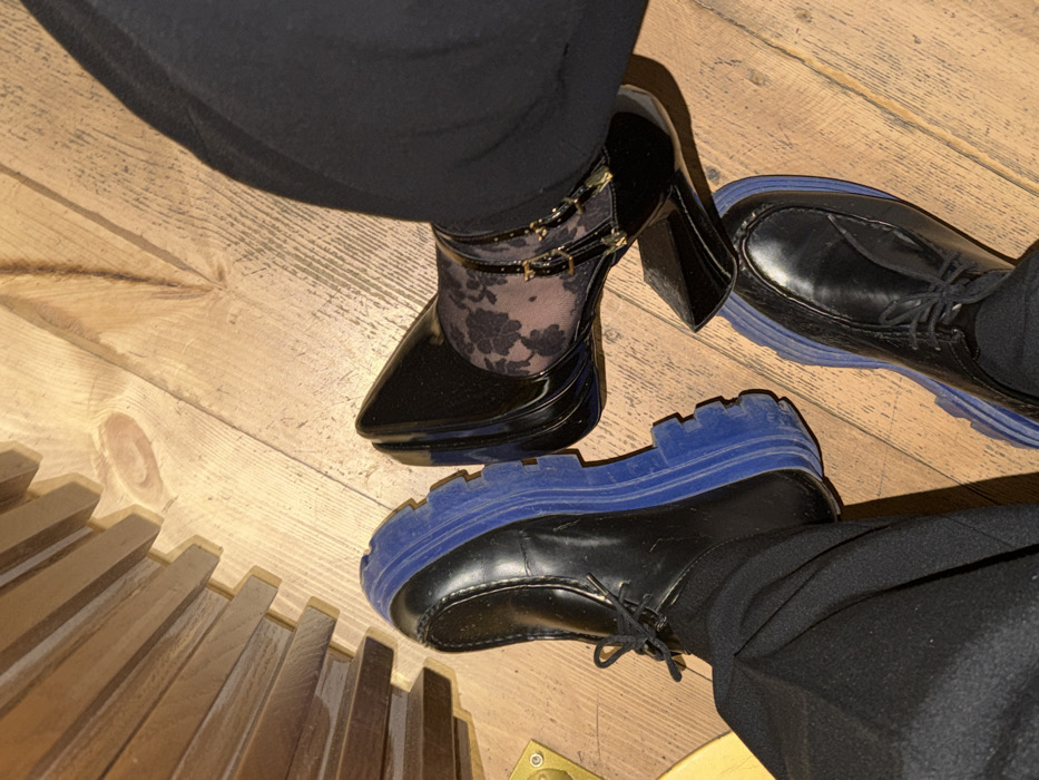 The image displays a ground-level, overhead perspective of two individuals' lower legs and feet on a light brown wooden floor, located in Yerevan, Armenia.
One individual, positioned towards the lower left, wears black trousers and black lace-up shoes with distinctive thick, ridged blue soles. The second individual, positioned towards the upper right, wears black trousers, sheer hosiery with a black floral or lace pattern, and black patent leather high-heeled shoes. These shoes feature pointed toes and two slender straps across the instep, each adorned with small golden buckle-like elements. The toe of the high-heeled shoe rests against the blue sole of the adjacent lace-up shoe.
The floor is constructed from wooden planks exhibiting natural grain patterns. To the far left, a partial view of a dark brown wooden structure with horizontal slat detailing is visible, along with a small, circular brass-colored fixture near its base.