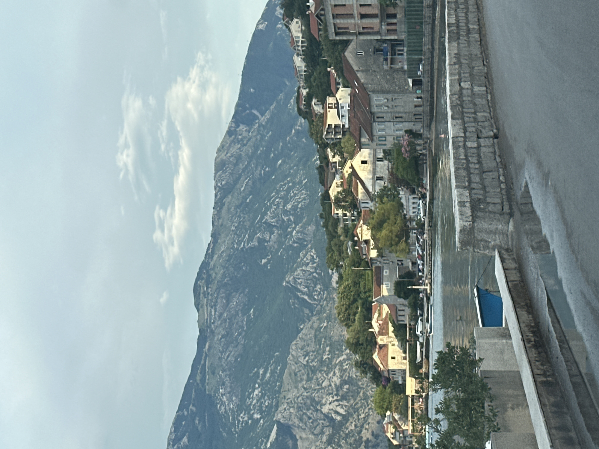 The image shows a view of the town of Prčanj, Montenegro, situated on a hillside overlooking a body of water. The foreground features a paved road and a stone wall. Beyond the wall, a body of water reflects the light, with a small blue boat visible near the shore. Houses with red tile roofs are clustered on the hillside, interspersed with trees and greenery. In the background, a large, rugged mountain range rises steeply, its rocky slopes covered in vegetation. The sky is partly cloudy, with a soft, diffused light suggesting either early morning or late afternoon. No people are clearly discernible in the scene.