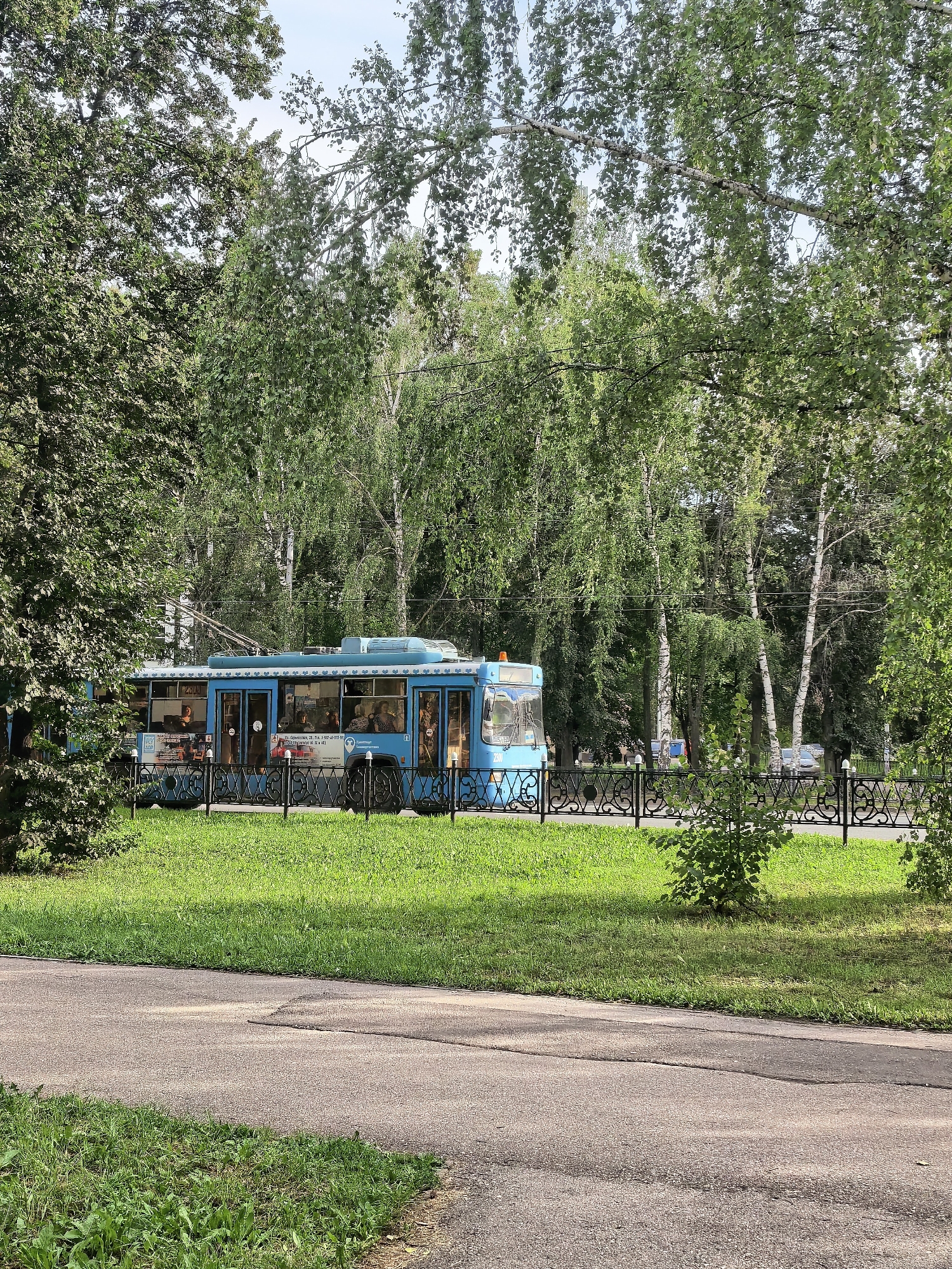 A bright blue trolleybus with advertising on its side travels along a paved road bordering a grassy area. The trolleybus has multiple windows and appears to be carrying passengers. Behind the trolleybus is a black decorative metal fence, and beyond that, a densely wooded area with tall birch and other deciduous trees. The sky is a pale blue with some scattered white clouds. In the foreground, a paved pathway runs diagonally from the bottom left corner towards the center right of the frame, with grass on either side of it. The image appears to be taken on a clear, sunny day. The specific location is Komsomolsky Park in Sterlitamak, Russia, near the intersection of Komsomolskaya Street and Oktyabrya Avenue.