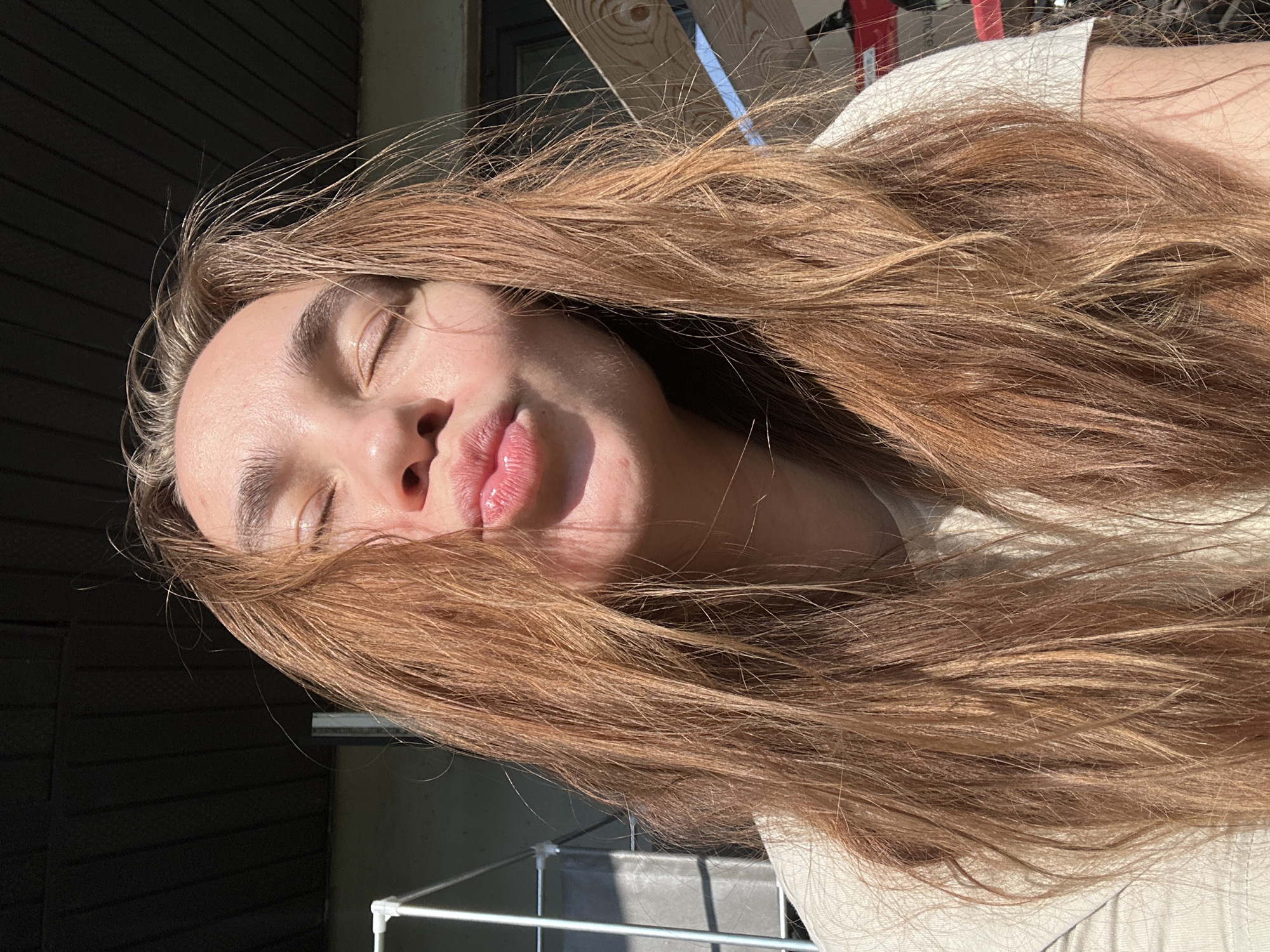 A young woman is lying with her head tilted to the right, eyes closed and lips pursed in a kiss. She has long, light brown hair. The scene is outdoors, with sunlight shining on her face and hair. A white metal railing is partially visible in the lower left corner, and a dark-colored wall is in the background. The location, Kharnavy, Russia, is not discernible from the image.