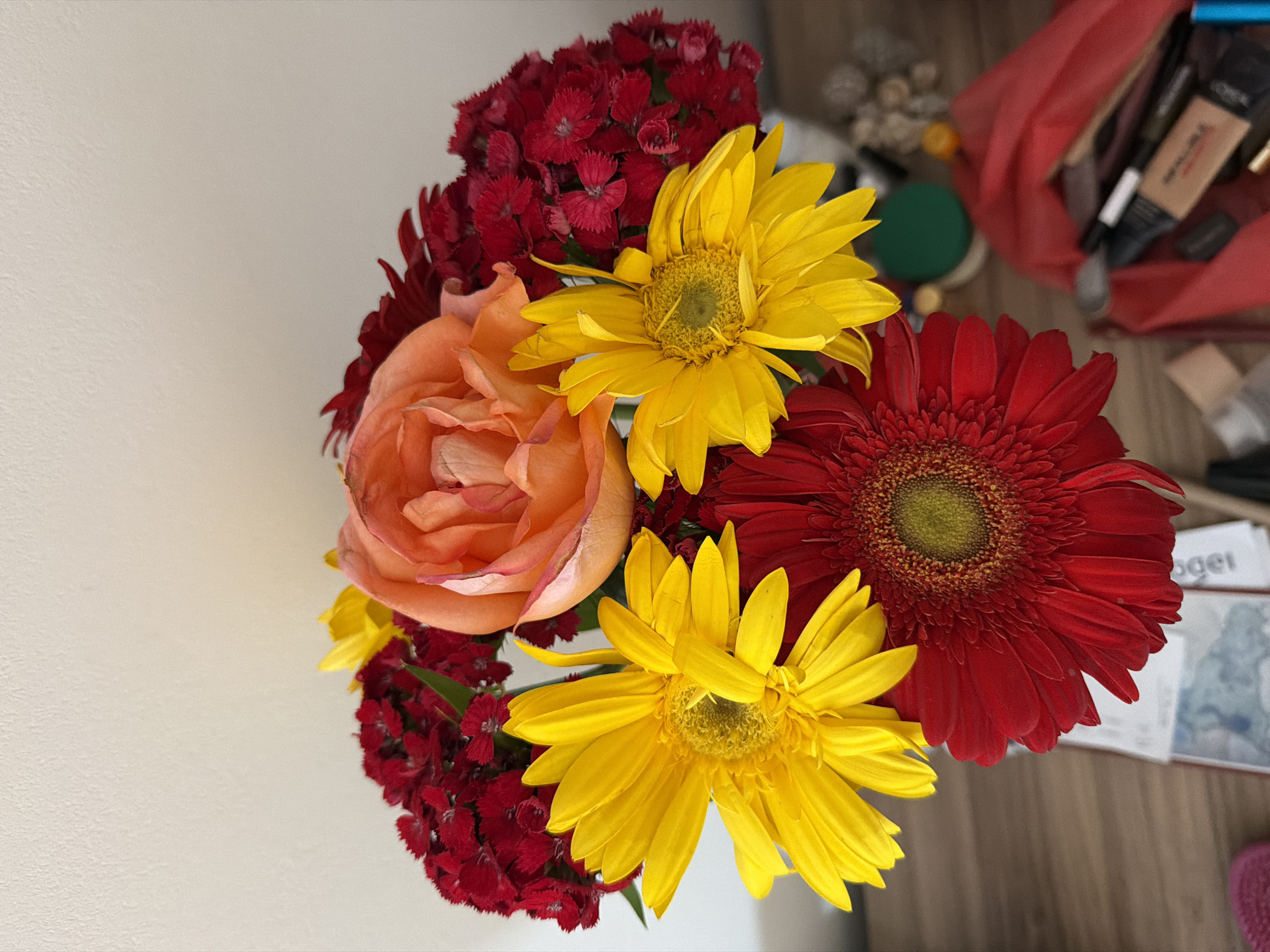 A still life image shows a bouquet of flowers in bright colors. The flowers include red carnations, a peach-colored rose, two yellow sunflowers, and a red gerbera daisy. The flowers are arranged closely together, filling the frame.
In the background, a red cloth bag is visible with various items scattered around it, including what appear to be makeup products and containers. There are also pieces of paper with blue smudges and printed words on them.
Without additional information from the image, it is not possible to verify if it was taken in Catania, Italy or specify actions, interactions, or the nature of the scattered items.