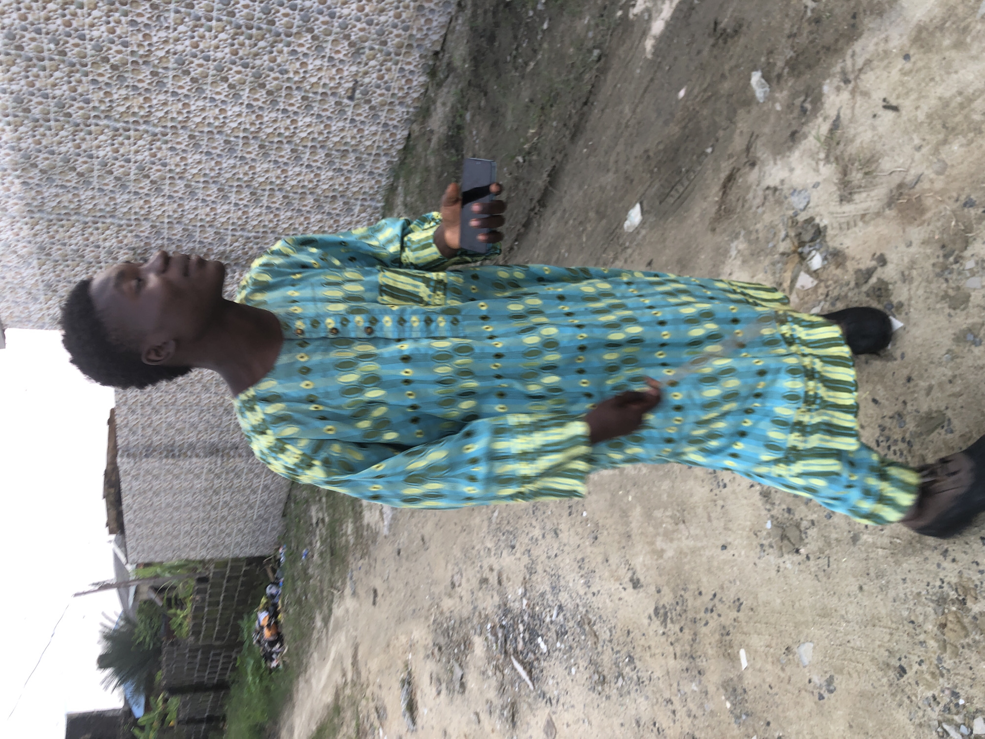 A young Black male, with short dark hair, stands on a dirt road in Ado, Nigeria, looking to his right. He is wearing a long, blue and green patterned robe with vertical stripes and circular patterns. He holds a dark-colored smartphone in his right hand. The background features a textured, light-colored wall on the right and a brick wall with overgrown vegetation to the left. Scattered debris is visible on the road and to the left of the road. The lighting suggests it is daytime, possibly overcast.