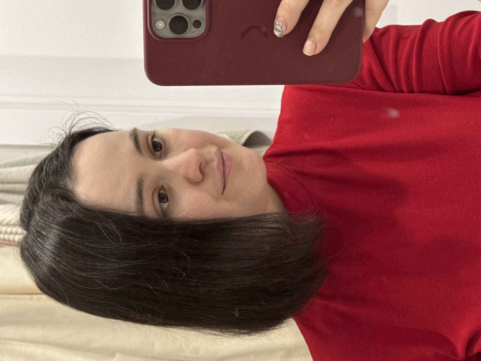 A woman with dark hair is taking a selfie. She is wearing a red shirt. The phone she is using is in a dark red case. Her nail is painted with a design. She is indoors. No location information for Izhevsk, Russia can be inferred.
