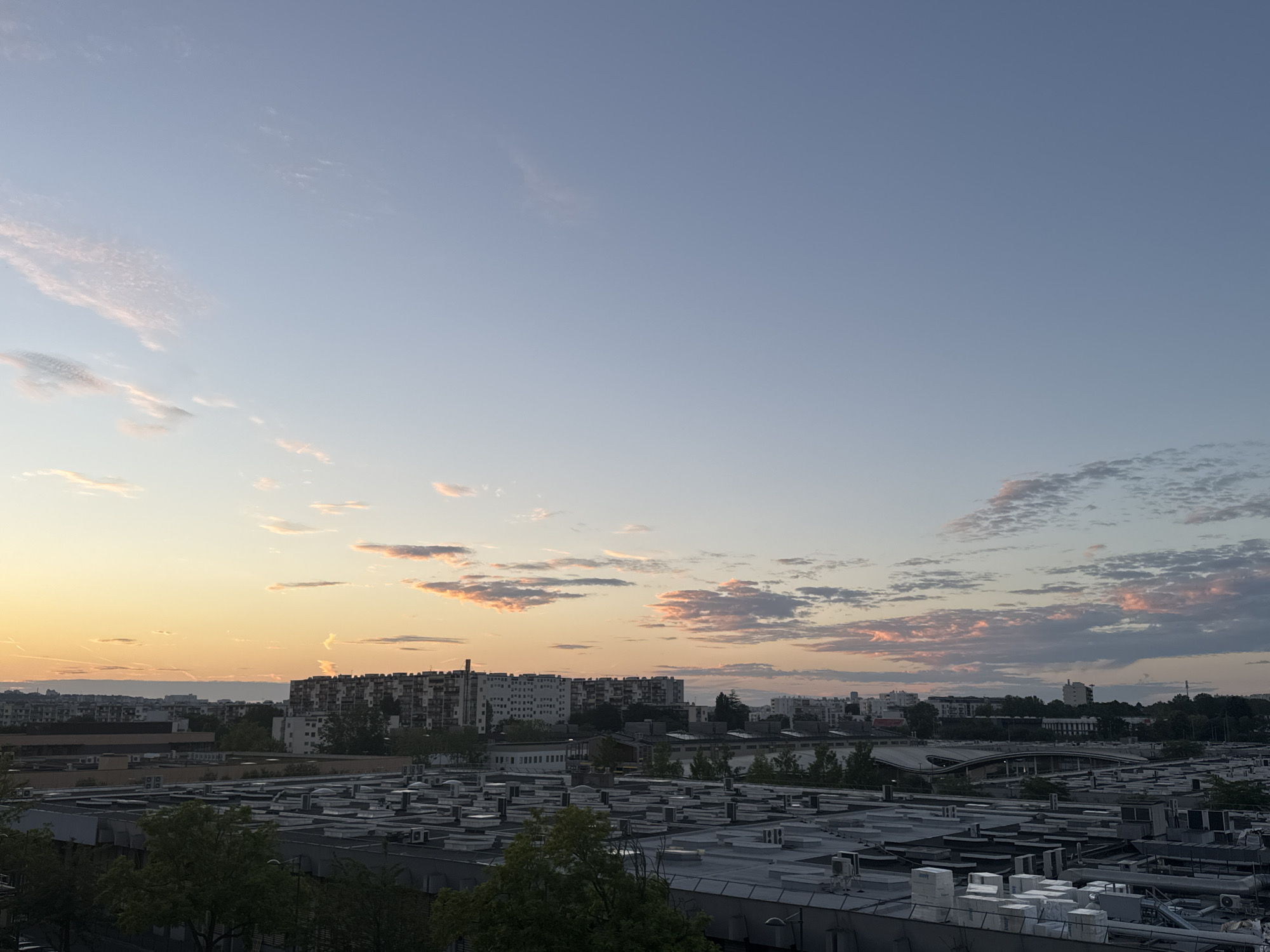 This is an aerial photograph of a cityscape at dusk. The sky is a gradient of pale blue and soft orange, with scattered clouds tinged with pink and white. The horizon is lined with a dense urban landscape, featuring numerous residential buildings and industrial structures. In the foreground, the rooftops of several low-rise buildings are visible, covered in HVAC units and other equipment. Trees with green foliage are interspersed among the buildings, suggesting a relatively green urban environment. The overall impression is that of a city at the end of the day, with the fading light casting a warm glow over the scene.