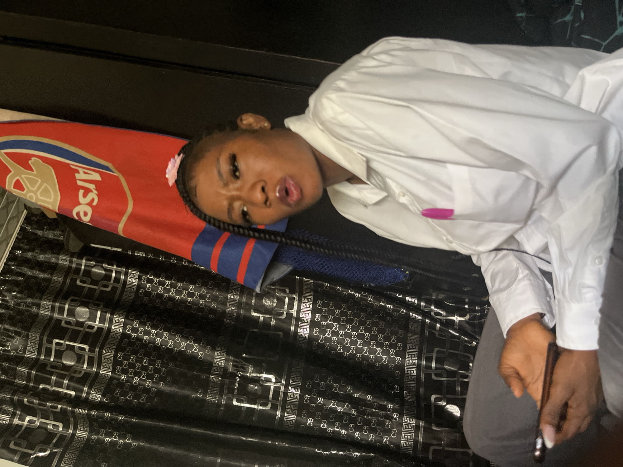 In a close-up, low-angle shot, a young Black woman is lying down, her head resting on a surface. She is wearing a white button-down shirt and grey pants. Her hair is styled in braids, with a small pink accessory visible near her forehead. Her eyes are open, and her mouth is slightly parted, conveying a relaxed or perhaps playful expression.
Behind her, a red and blue object with the "Arsenal" logo is visible, suggesting a connection to the football club. The surface she is lying on appears to be covered with a black fabric featuring a repeating silver geometric pattern, possibly a blanket or upholstery. The overall setting seems to be indoors, with dark, out-of-focus elements in the background.
The lighting is moderate, casting soft shadows and highlighting the textures of the fabric and the woman's clothing. There are no clear indicators of the time of day or weather conditions. The scene is static, with the woman posed rather than actively engaged in an event. The visible text is "Arsenal" on the red and blue item. The presence of the Arsenal logo might suggest a fan's personal space or a themed environment.