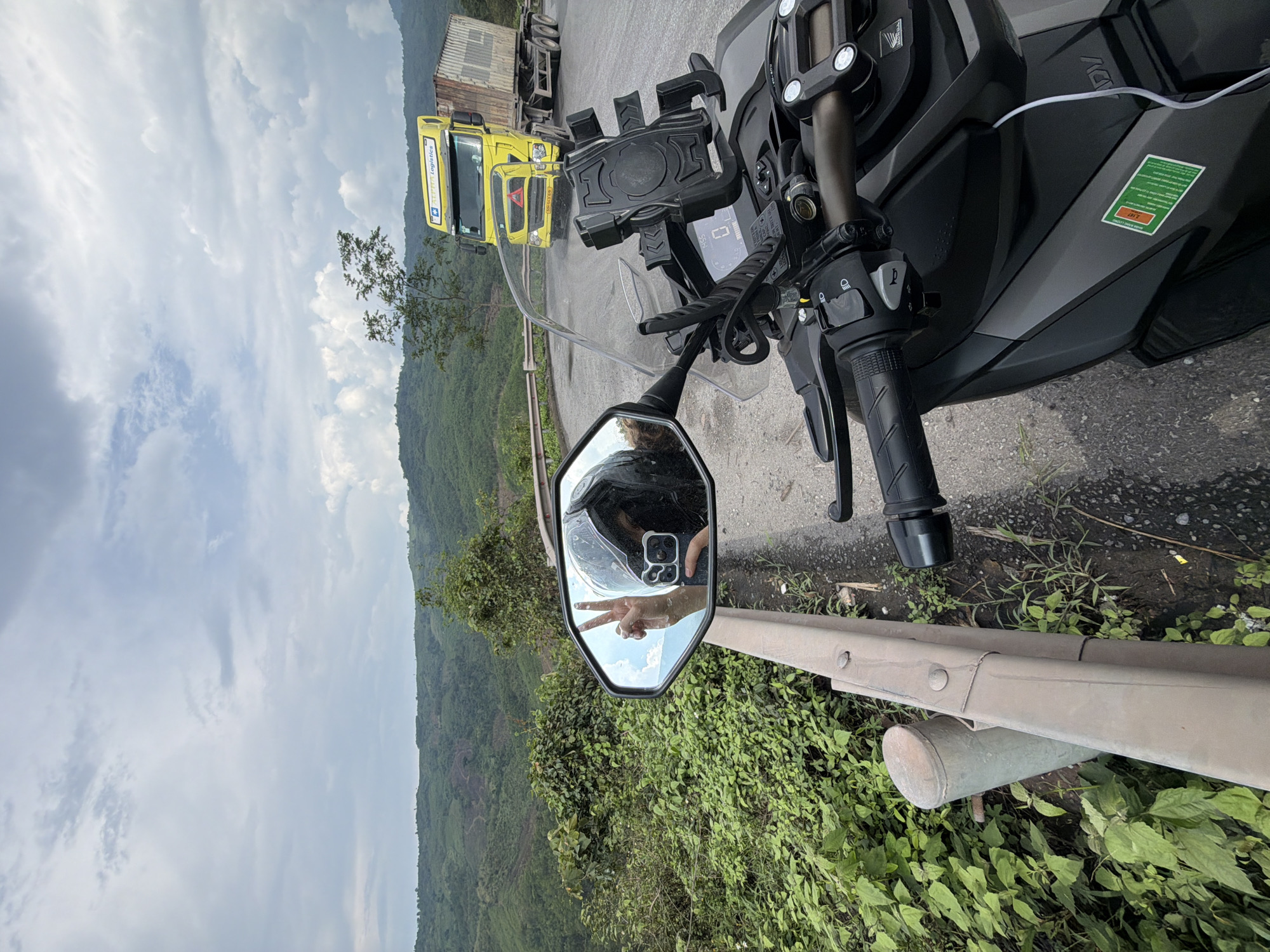 The image depicts a motorcycle parked on the side of a road in La Lay Commune, Vietnam, with a view of a lush, hilly landscape. In the foreground, the motorcycle's handlebar, instrument panel, and rearview mirror are visible. Reflected in the mirror is a hand making a peace sign and a helmet. A guardrail runs along the edge of the road, separating it from the vegetation. Beyond the guardrail, dense greenery covers the slopes. Further in the distance, the hills gradually fade into the background. In the upper-left corner, part of a yellow truck and a trailer are visible on the road. The sky is overcast with scattered clouds.