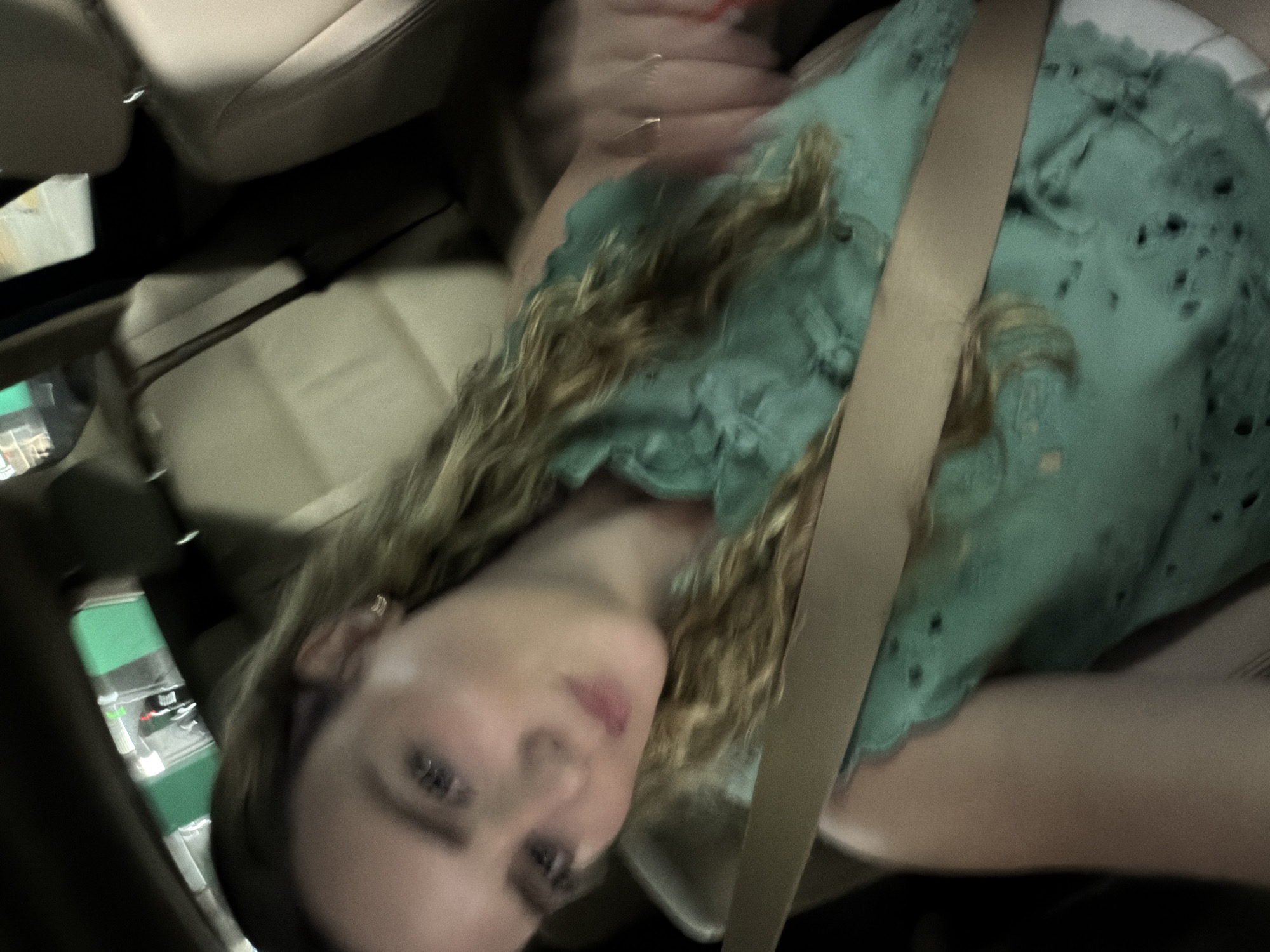 A young woman is seated in the passenger seat of a car, wearing a green shirt and a seatbelt. The interior of the car is light beige. Out the window, green and white structures are partially visible. The location is specified as Miami, United States of America, although there is no indication in the photo to corroborate this information.