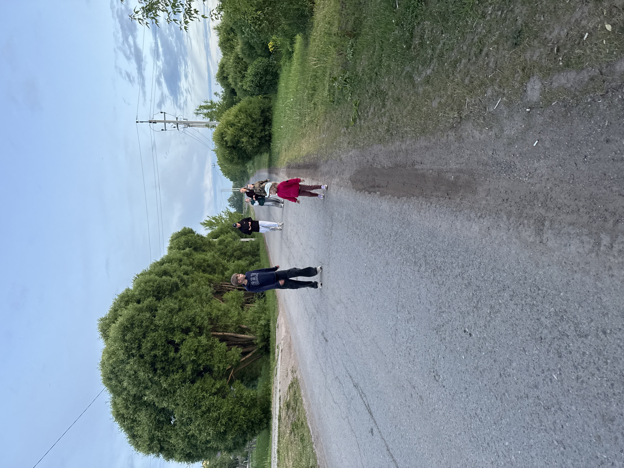 The image captures a road in Горный, Russia, with a group of people walking along it. The road surface appears to be asphalt. The terrain around the road is characterized by greenery, including trees and grass. Utility poles with power lines are visible. In the foreground, a person is standing and facing the viewer. Further down the road, several individuals are seen walking away from the camera. The sky is overcast.