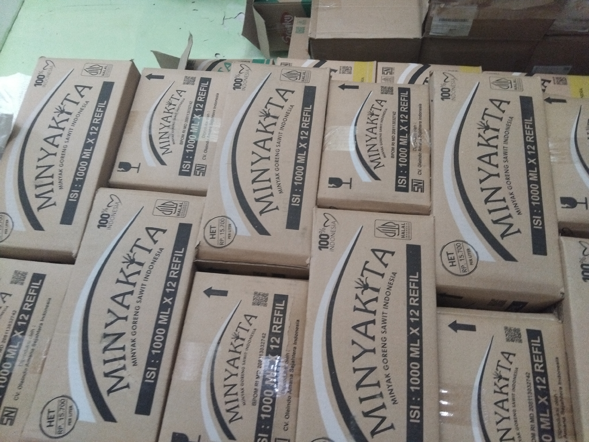 A stack of cardboard boxes of "Minyakita" brand cooking oil is shown in a dimly lit storage area. The boxes are cream-colored with black text and graphics. Each box displays the brand name "Minyakita" in large, arched lettering, followed by "MINYAK GORENG SAWIT INDONESIA" (Indonesian Palm Cooking Oil). Below this, "ISI: 1000 ML X 12 REFIL" (Contents: 1000 ML X 12 Refills) is printed. Other markings include "100% INDONESIA", a Halal certification logo, SNI (Indonesian National Standard) logo, and a price indication "HET RP. 15.700 PER LITER". The boxes are arranged in a somewhat disorganized manner, suggesting a commercial storage environment. The background is out of focus, but appears to be a wall or other storage structures. This scene is located in the city of Berbek, Indonesia.