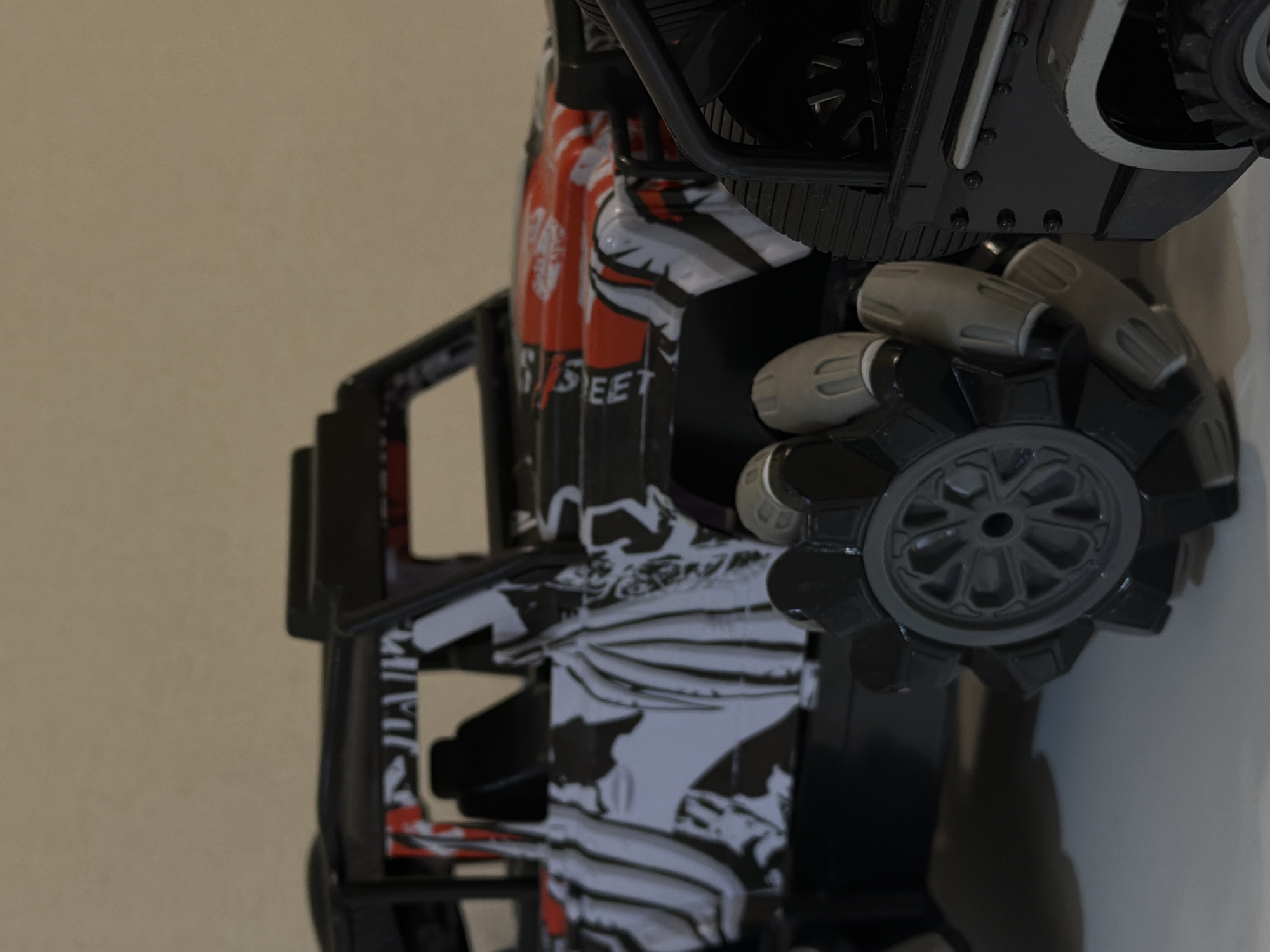 This close-up image captures a toy remote control vehicle, likely an RC car or truck, viewed from a side angle with a slight tilt. The primary subject is the toy, which displays a distinctive aesthetic. Its body is adorned with a dynamic graphic pattern in black, white, and red. Notable design elements include a robust black frame or roll cage that surrounds the main body.
A prominent feature of the toy is its unusual wheels, which appear to be multi-directional or mecanum wheels, characterized by multiple rollers set around the circumference. These grey-colored wheels suggest advanced maneuverability. One such wheel is clearly visible in the foreground, showing its intricate design.
On the side of the vehicle's body, the word "STREET" is distinctly visible in white capital letters against a dark background, part of the overall graphic design. Other abstract patterns and lines are also part of the bodywork.
The vehicle is presented against a plain, light brown or beige background, suggesting an indoor setting, possibly a room with a simple wall. No specific activity is depicted; the image is a static shot of the toy. The lighting is diffused, highlighting the textures and colors of the vehicle without harsh shadows, and there are no discernible location cues in the image that correspond to the provided "Sofiivska Borshchahivka, Ukraine" context.