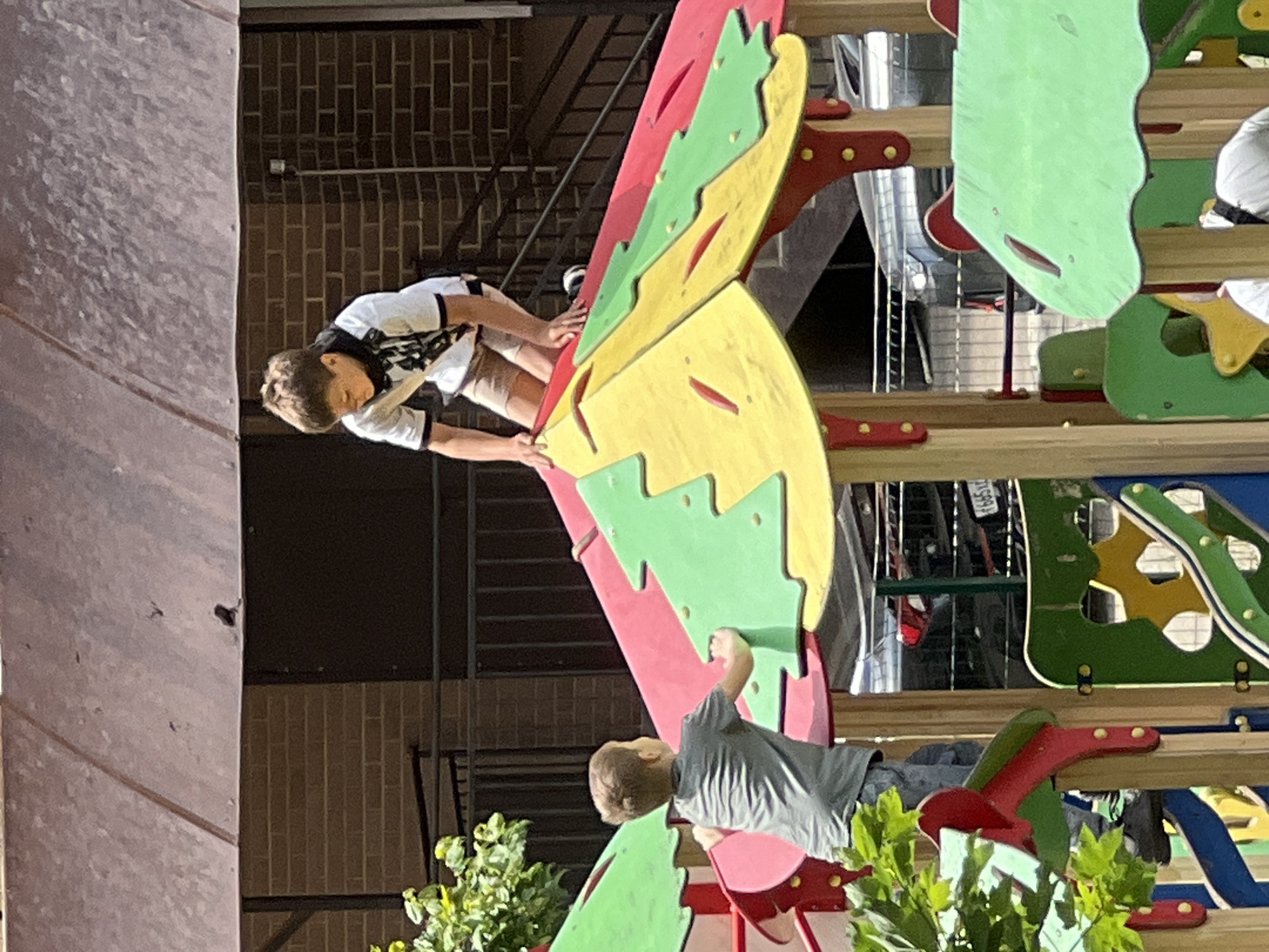 Two children are climbing on colorful playground equipment. The playground equipment is painted with red, green, and yellow paint. A bush is growing in front of the equipment. A brick building is behind the playground equipment. In the background, bicycles can be seen. The photograph was taken in Krasnodar, Russia.