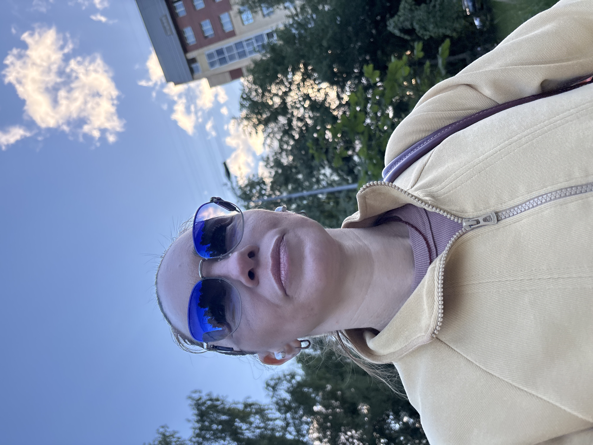 A woman wearing sunglasses and a light yellow jacket is standing outside. She has earbuds in her ears and a purple strap of a bag is visible on her shoulder. In the background, there are trees and a tall building. The sky above is blue with some white clouds. This scene appears to be in the city of Sinyavino, Russia.